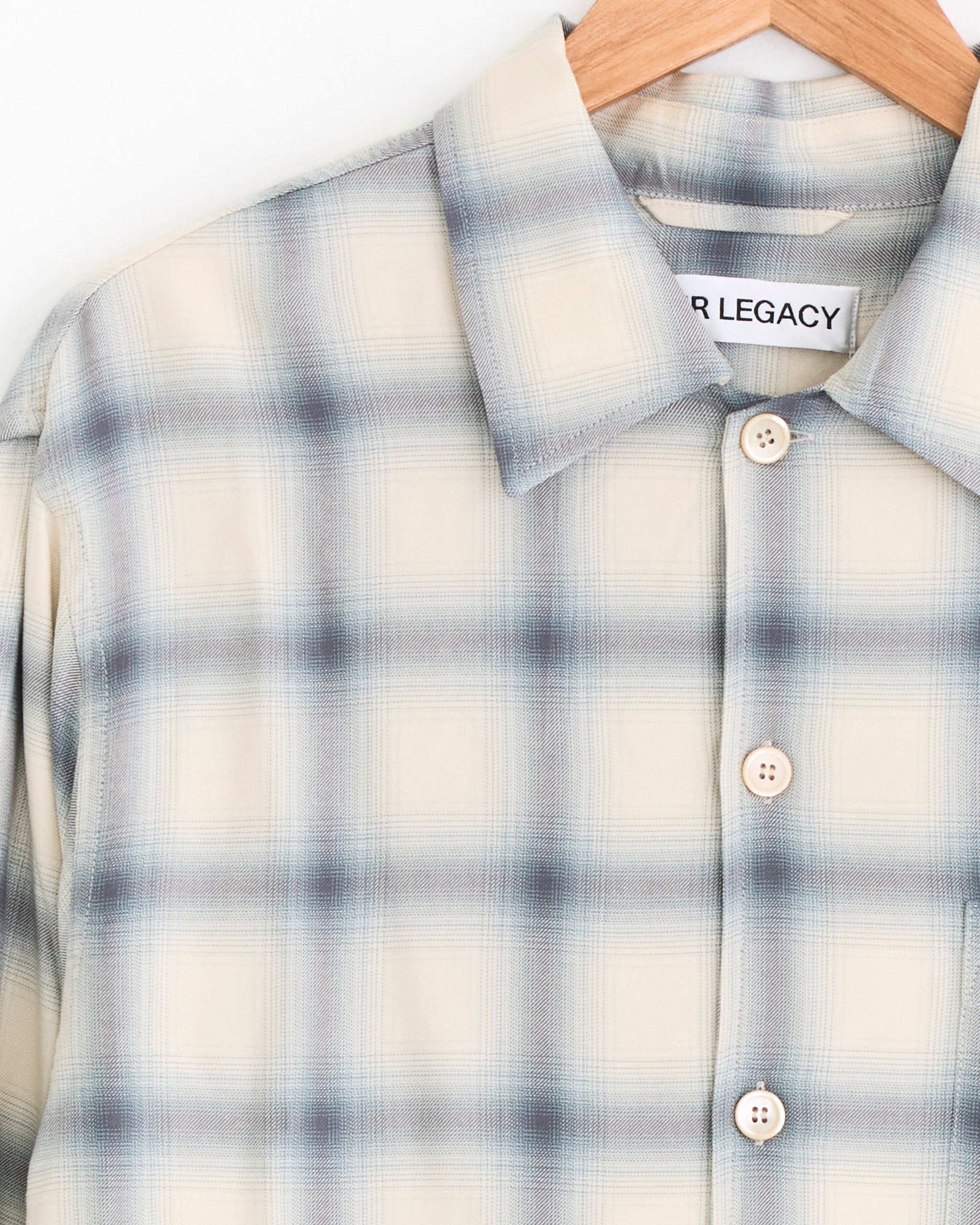 The Every Shirt in Sacramento Cowboy by Our Legacy is a beige and gray plaid button-up with a classic collar and white buttons, displayed on a wooden hanger. The label reads OUR LEGACY.