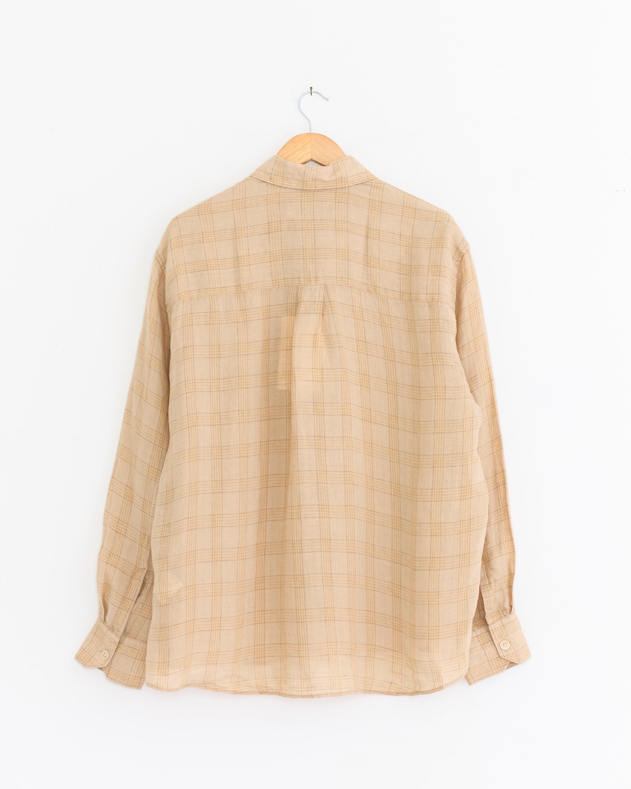 The Our Legacy Above Shirt in Vacay Check, featuring beige plaid linen and long sleeves, hangs on a wooden hanger against a plain white background, displayed from the back.