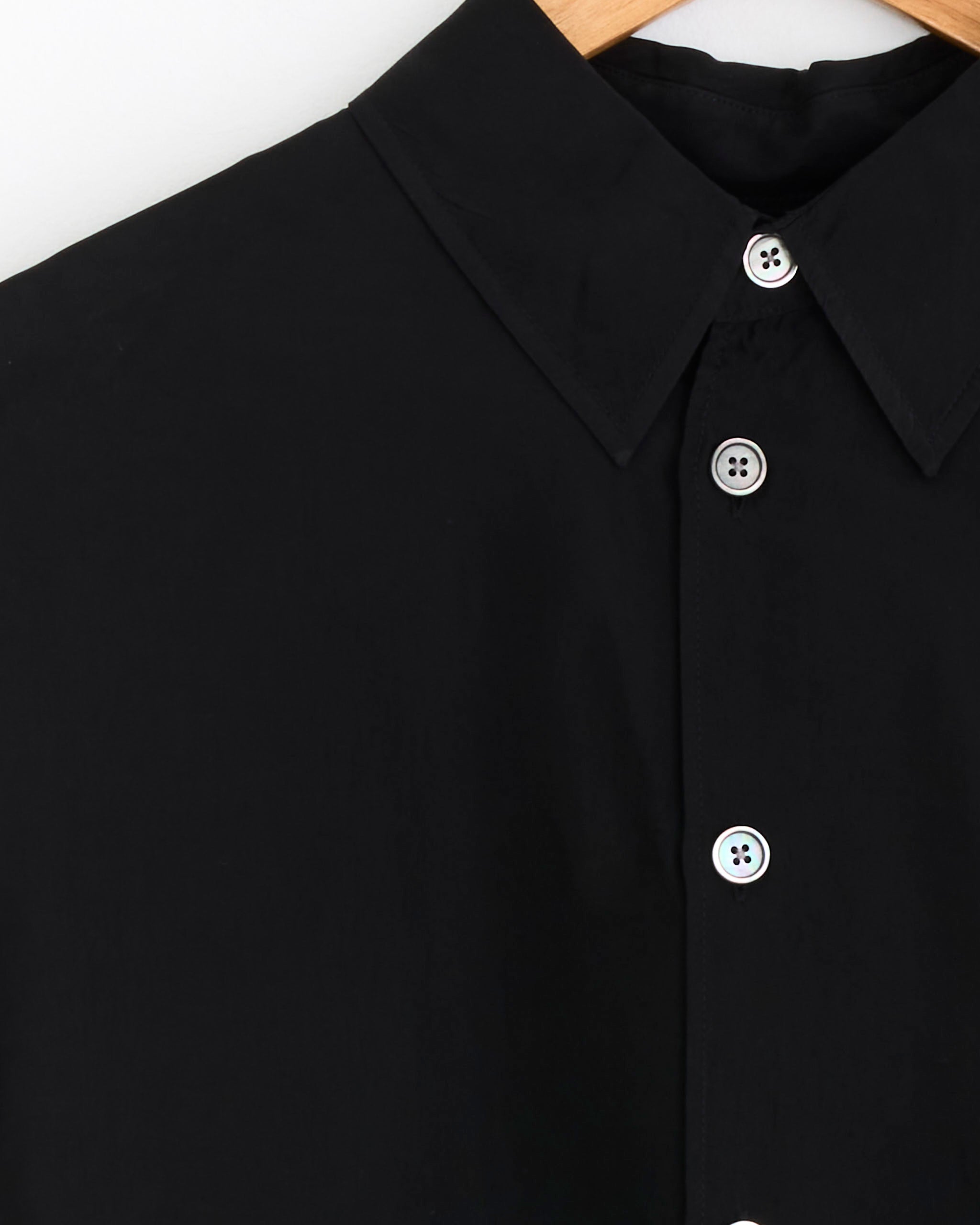 Close-up of the Our Legacy Beyond Shirt in Char Black, made from Japanese cupro fabric, featuring a collar, white buttons, and a wooden hanger visible at the top.