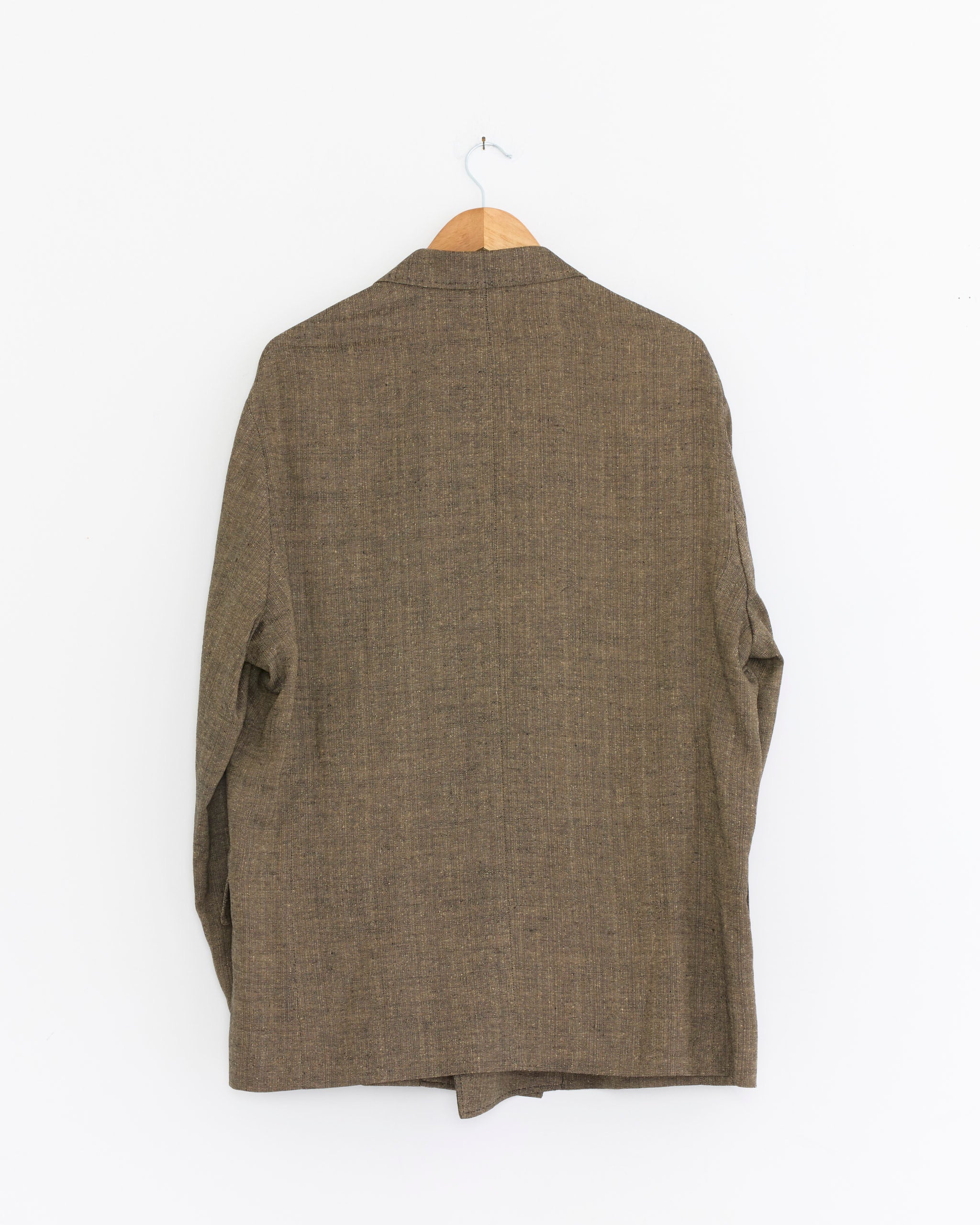 The Our Legacy Halt Blazer in Deep Husk, crafted from textured brown Italian linen, is displayed from the back on a wooden hanger against a plain white wall.
