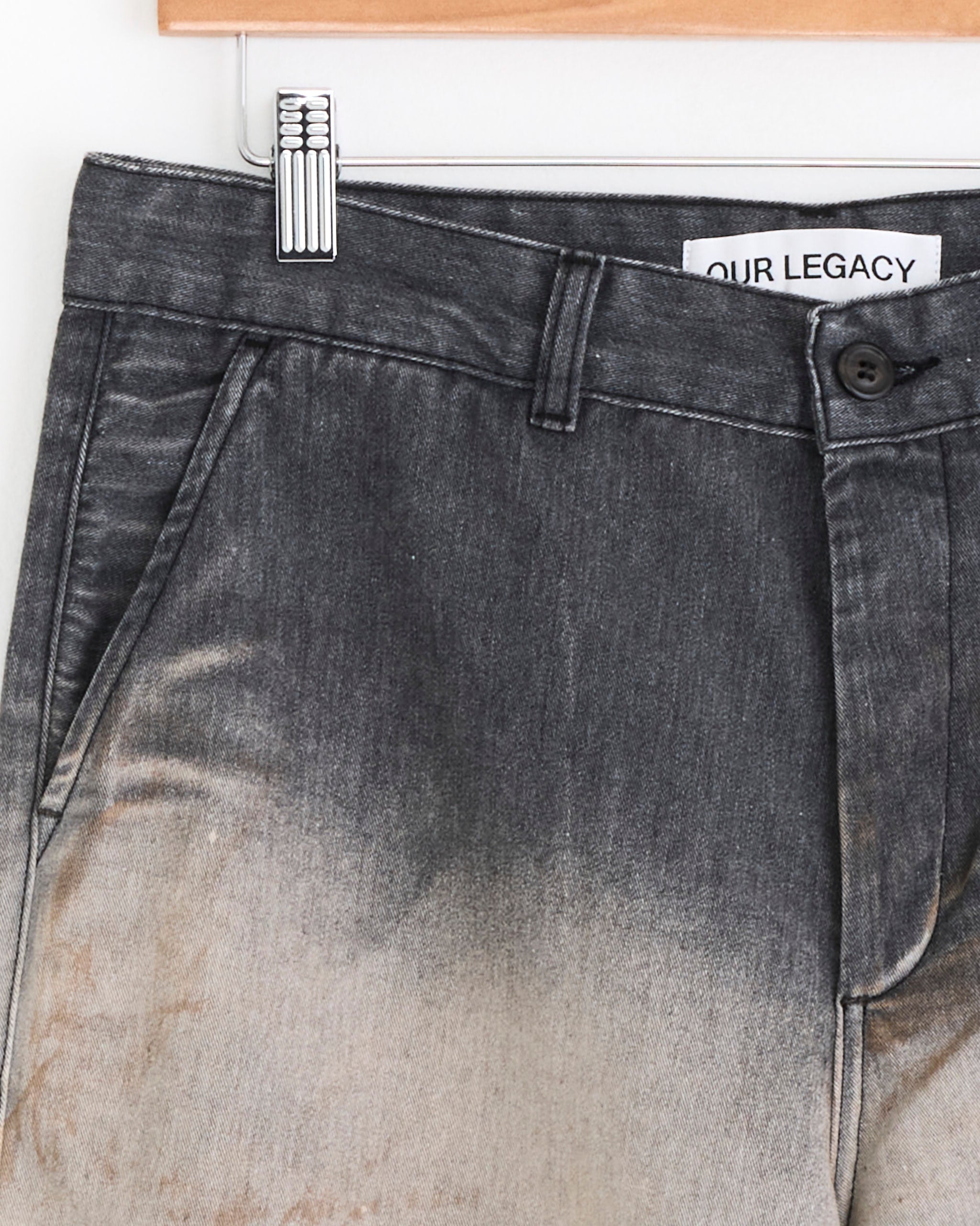 Close-up of Our Legacy Rig Chino in Octo Wash, featuring a faded black and beige vintage-wash denim, relaxed straight fit, visible waistband with button, belt loops, side pockets, and brand label on a wooden hanger.