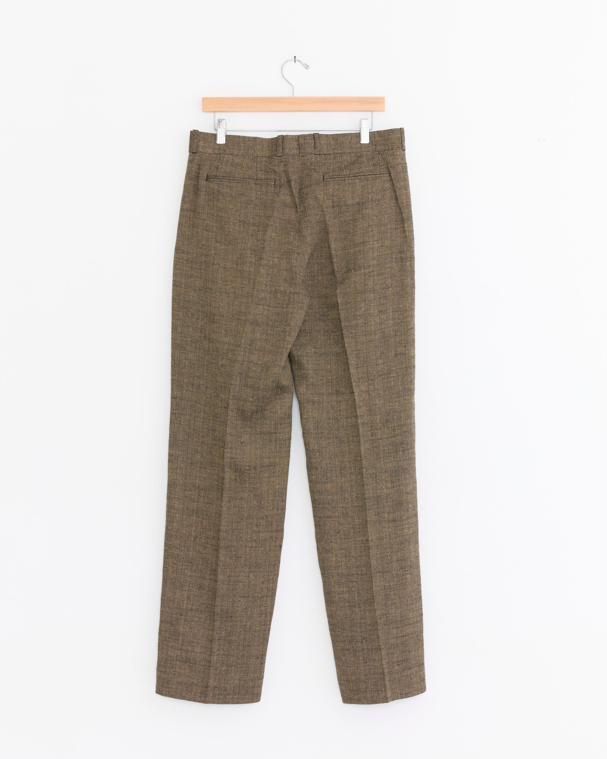 A pair of Our Legacy Slack Trouser in Deep Husk, made from an Italian linen silk cotton blend, hangs on a wooden hanger against a plain white background.