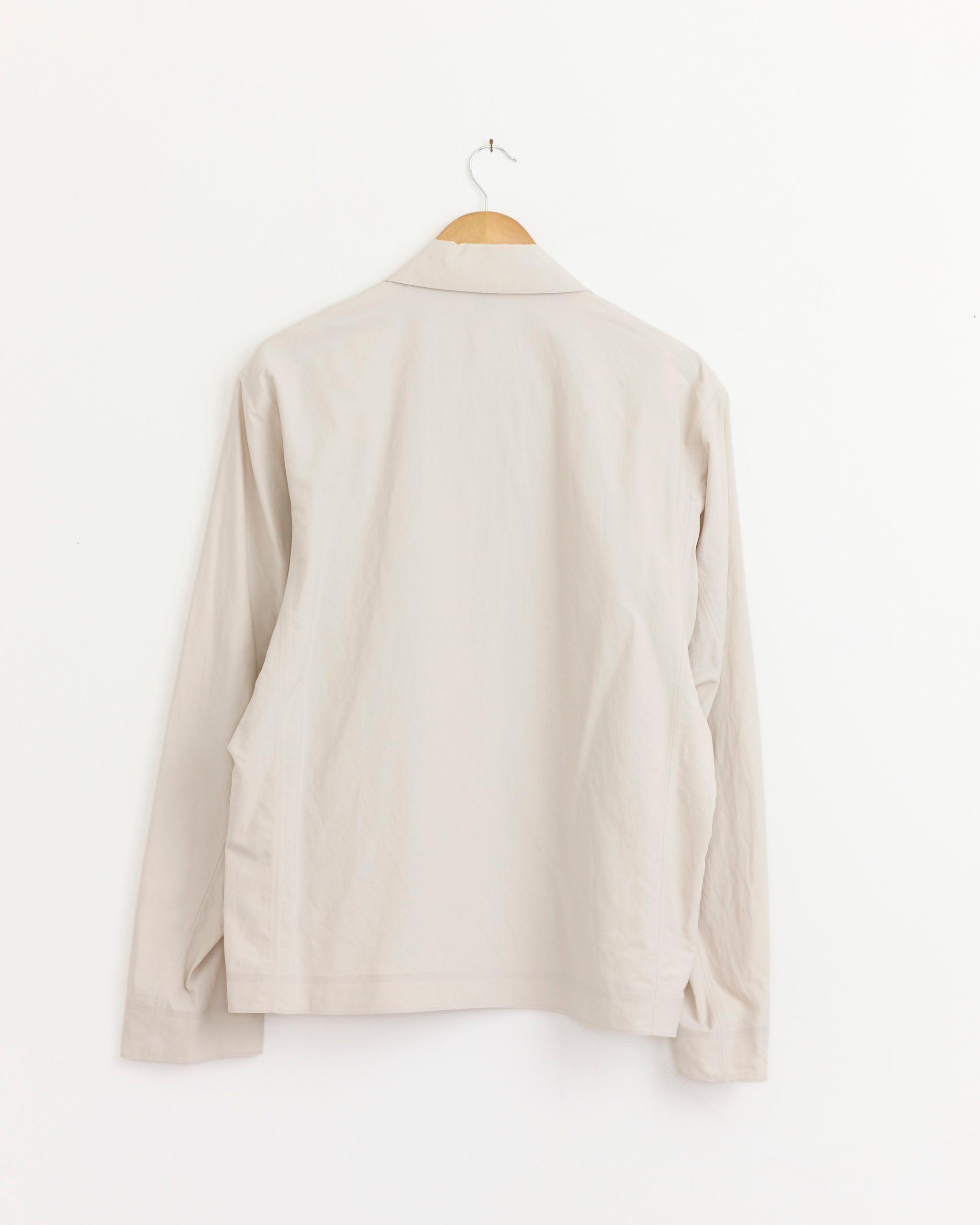 The Veilance Spere SL Jacket in Dark Cocoon is displayed on a wooden hanger against a plain white wall, shown from the back.