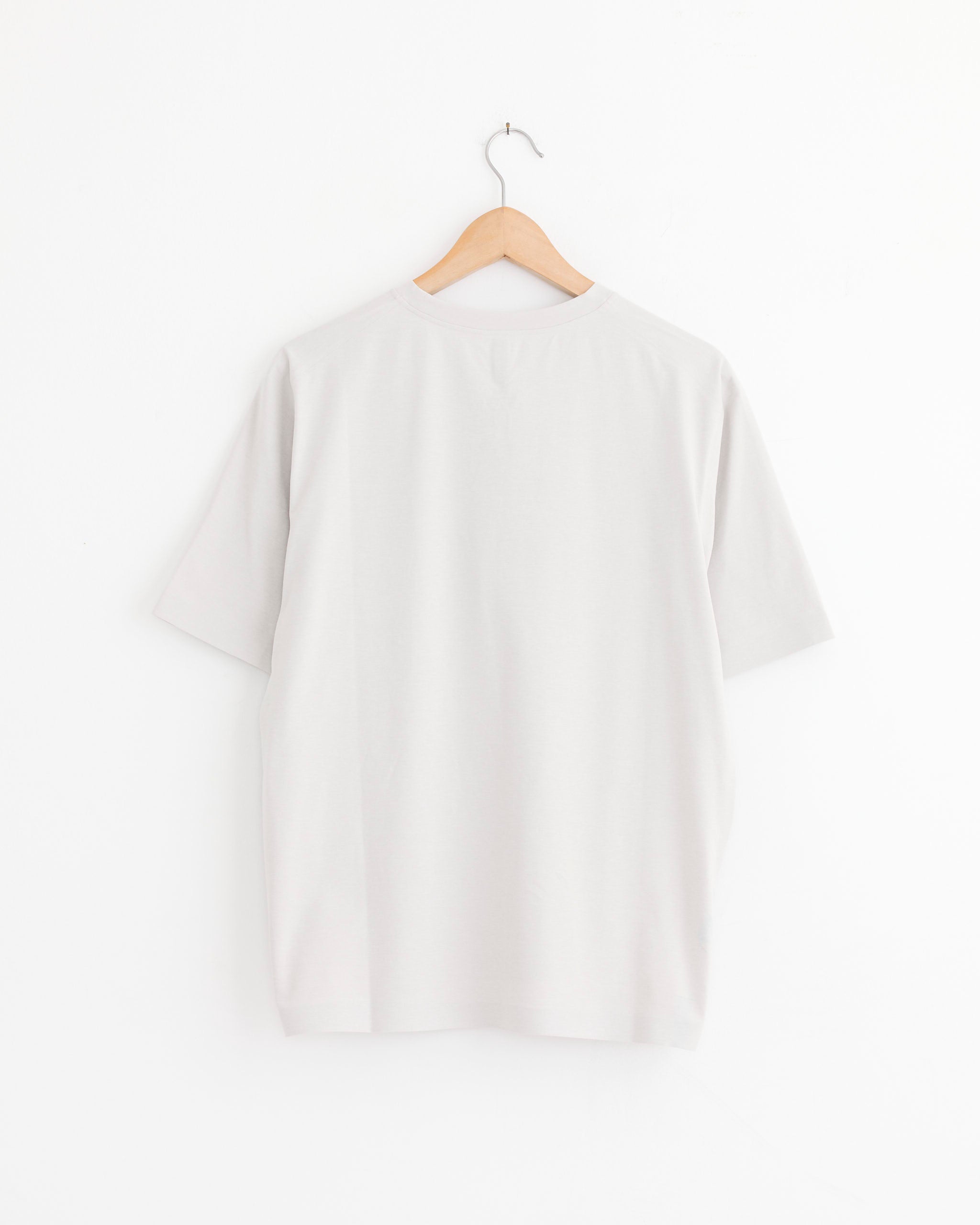 The Veilance Metron Tee in Dark Cocoon hangs on a wooden hanger against a white background, shown from the back—ideal for checking fit or discussing sizing via online chat.
