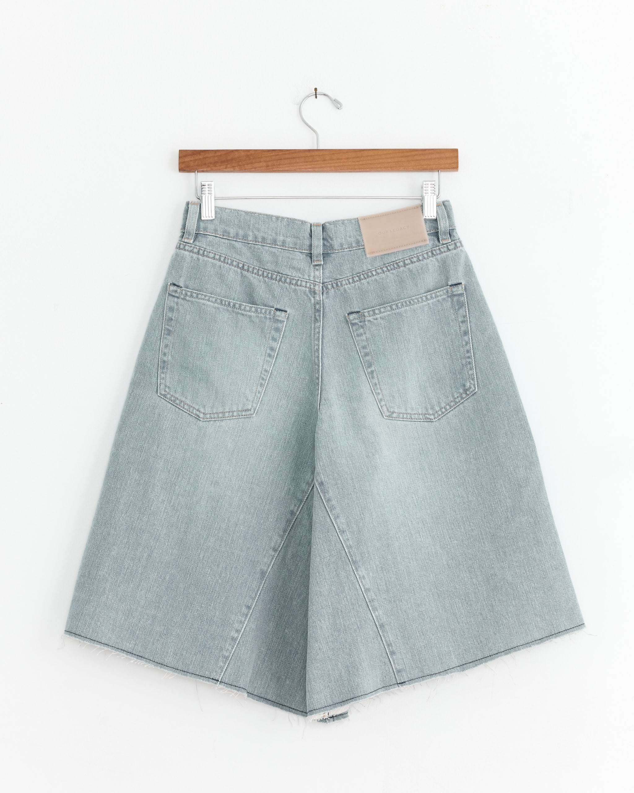 Our Legacy’s Midi Short in Pale Coast Wash—light blue, A-line denim shorts with raw hems, large back pockets, and belt loops—hangs on a wooden hanger against a white background.