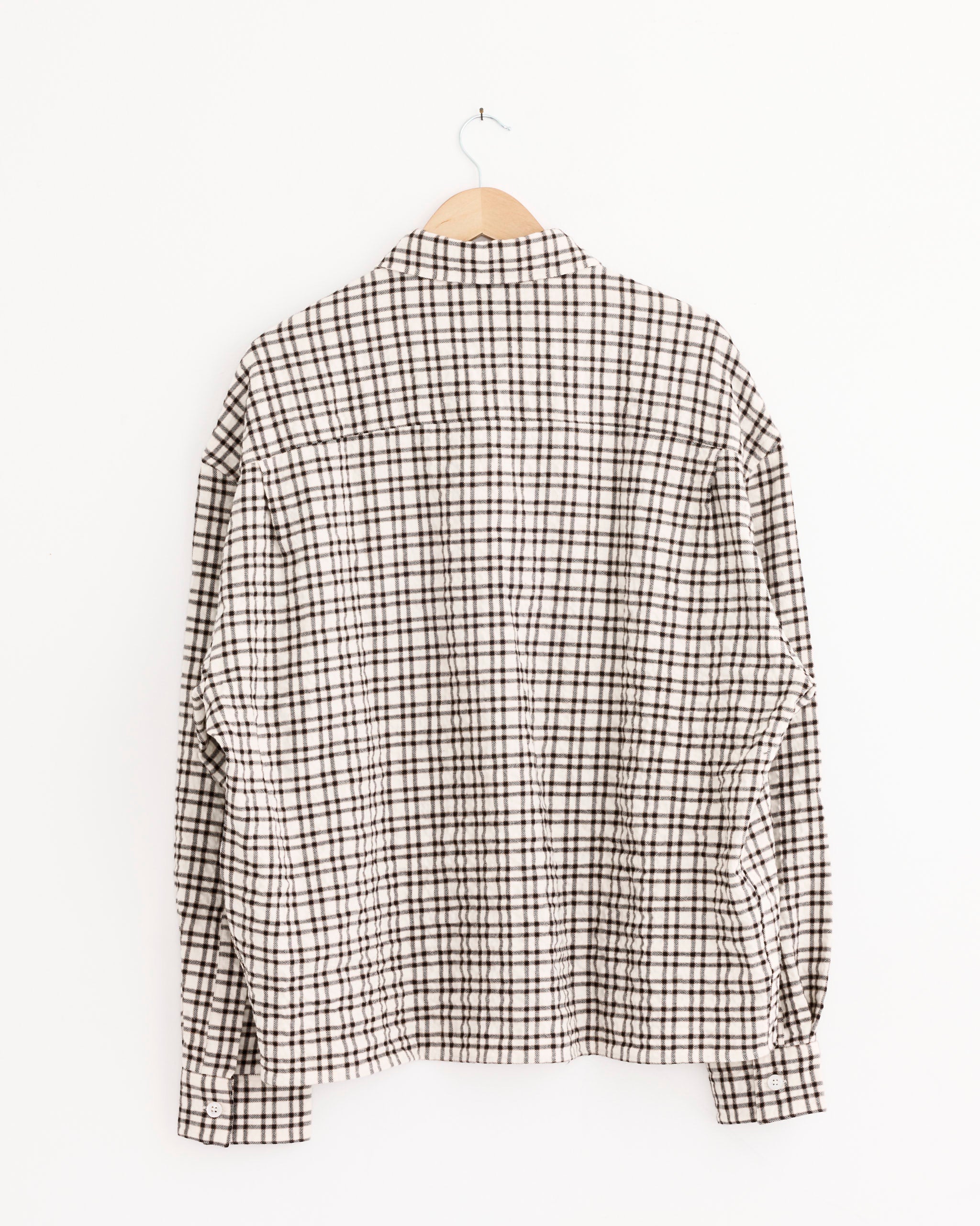 The Studio Nicholson Nostra Shirt in Canvas/Black Check, a long-sleeve button-up, is crafted from a wool-linen blend and hangs on a wooden hanger against a plain white background.
