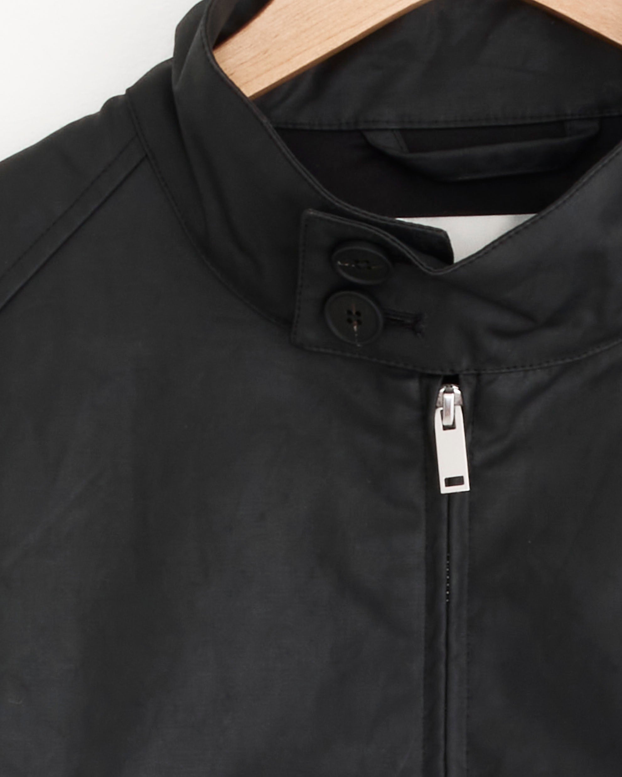 Close-up of the Studio Nicholson Over Jacket in Coal on a wooden hanger, showcasing its high collar with two buttons, visible silver zipper, and showerproof cotton-nylon fabric.