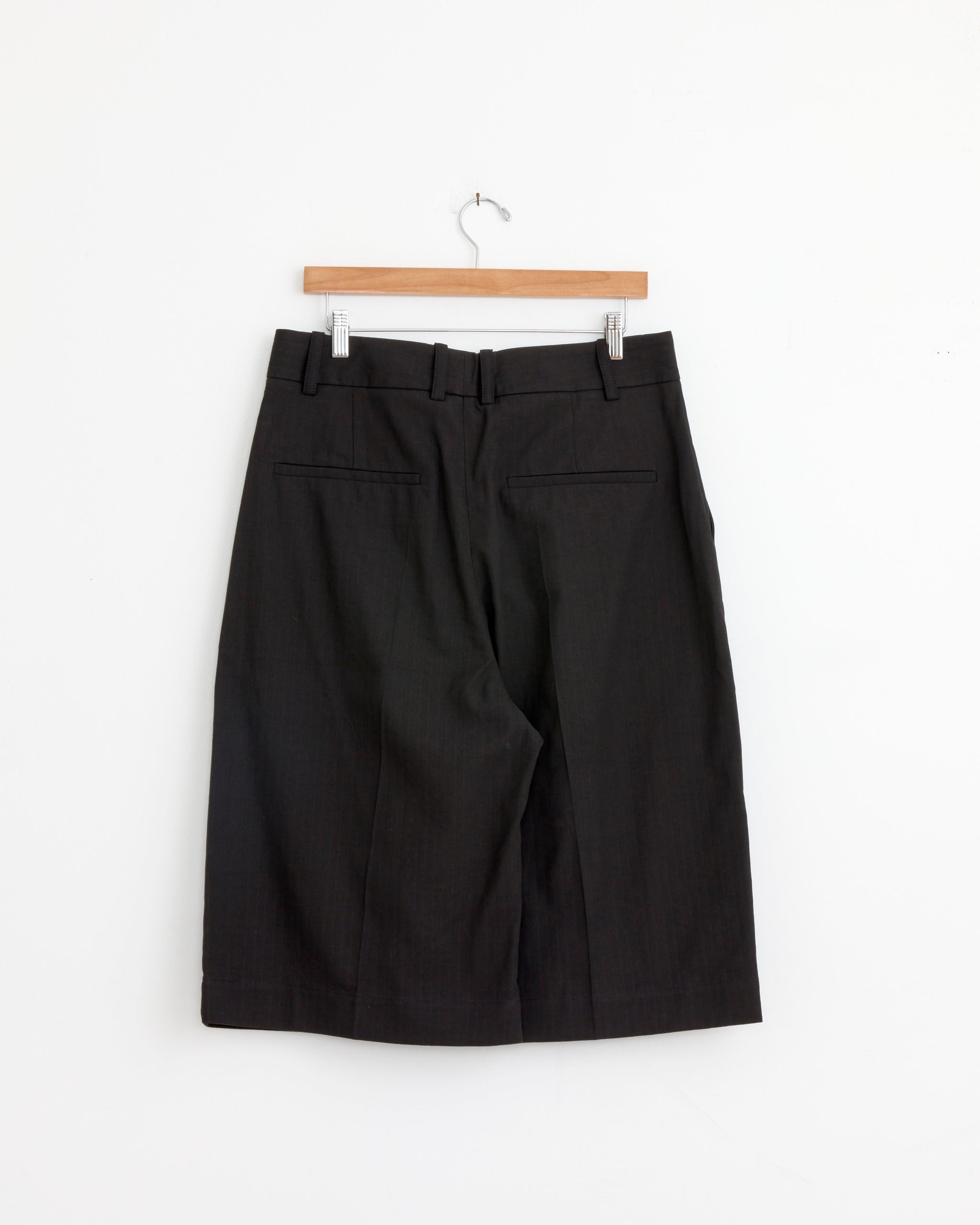 The Fowld Short in Black by Studio Nicholson, featuring an oversized tailored fit with belt loops and back welt pockets, hangs on a wooden hanger against a plain white background.