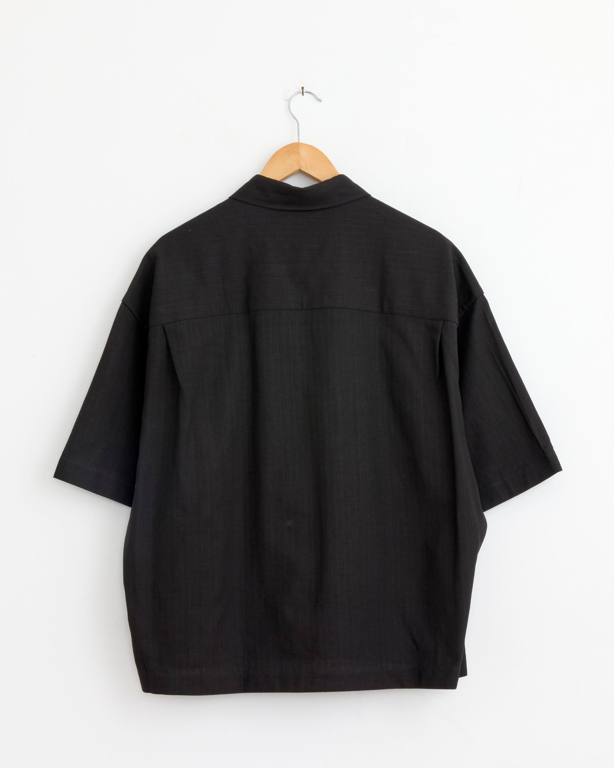 The Studio Nicholson Sanko Shirt in Black, an oversized, loose-fitting short-sleeve style, is displayed from the back on a wooden hanger against a plain white wall.