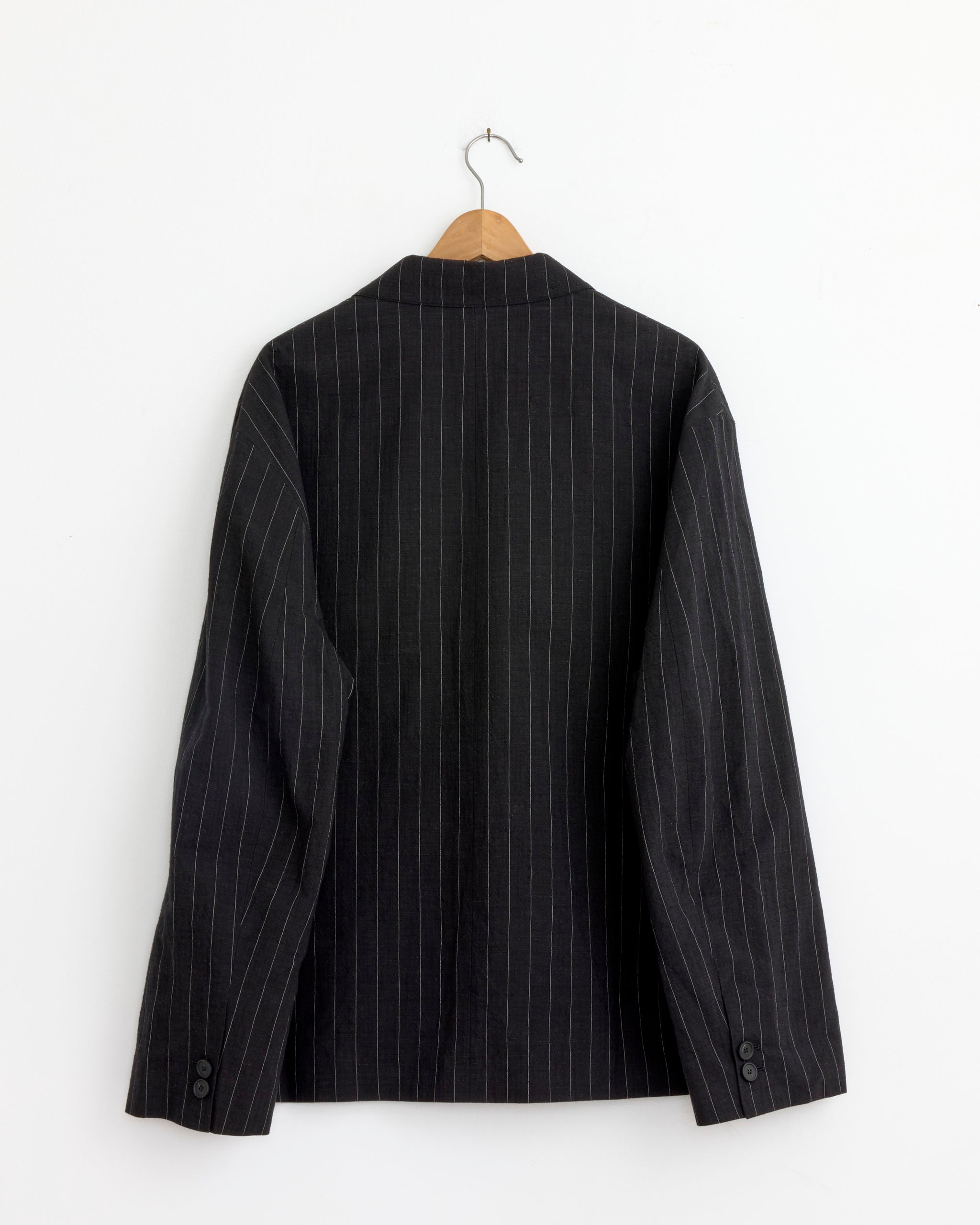 The Sorst Jacket in Black by Studio Nicholson, crafted from cotton linen with pinstripes, is displayed on a wooden hanger against a white wall, shown from the back with two buttons on each sleeve cuff.