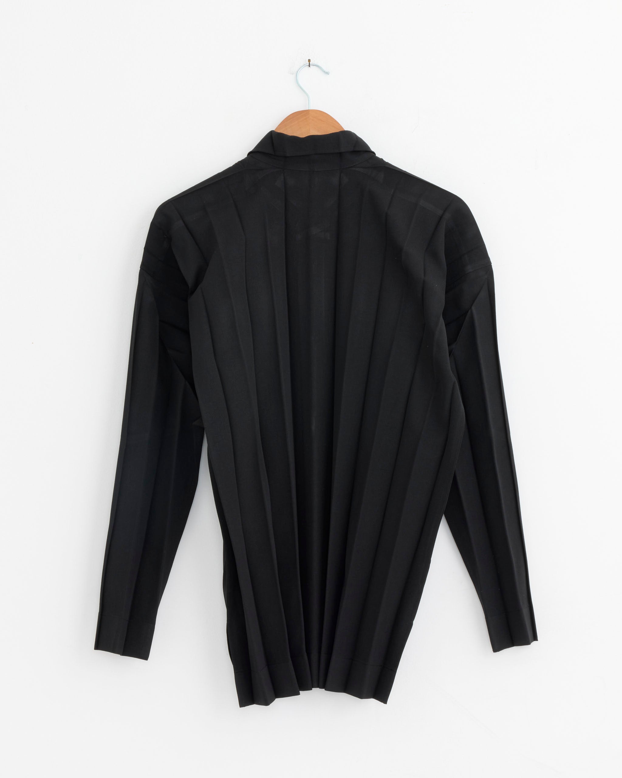 The Edge Ensemble Shirt in Black by Homme Plissé Issey Miyake, a long-sleeved pleated twill style, is shown from the back on a wooden hanger against a white background.