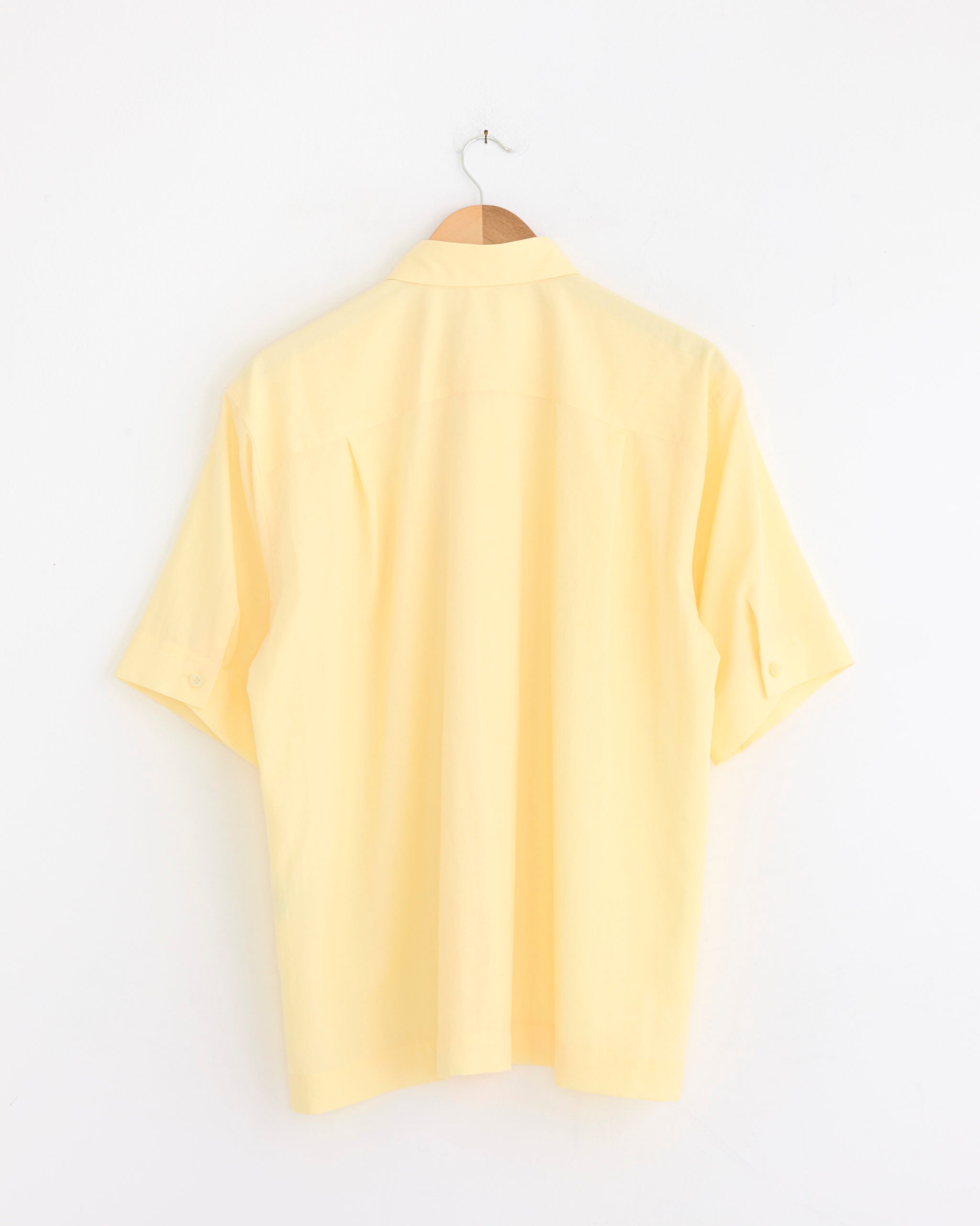 The PL Ramie Shirt in Yellow by Homme Plissé Issey Miyake, a wrinkle-resistant piece, is displayed on a wooden hanger against a white wall, with its back side visible.