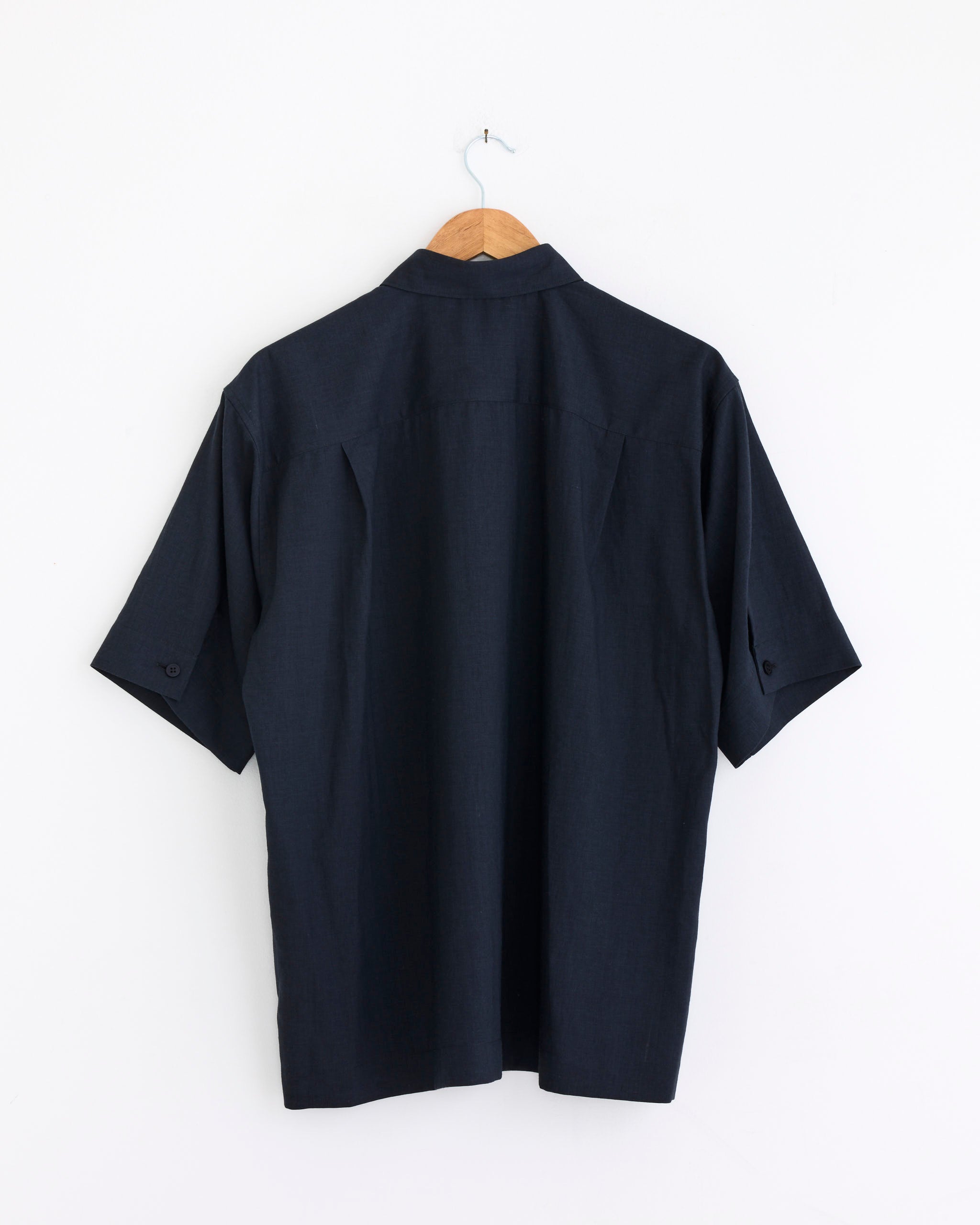 A short-sleeved Homme Plissé Issey Miyake PL Ramie Shirt in Charcoal hangs on a wooden hanger against a plain white wall, shown from the back—its wrinkle-resistant fabric keeps the look effortlessly stylish.