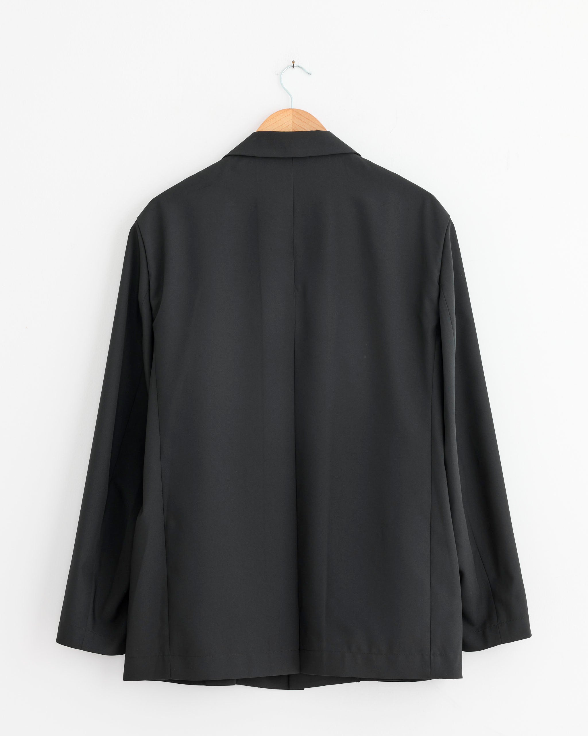 The IM MEN Wings Blazer in Black hangs on a wooden hanger against a white wall, displayed from the back.