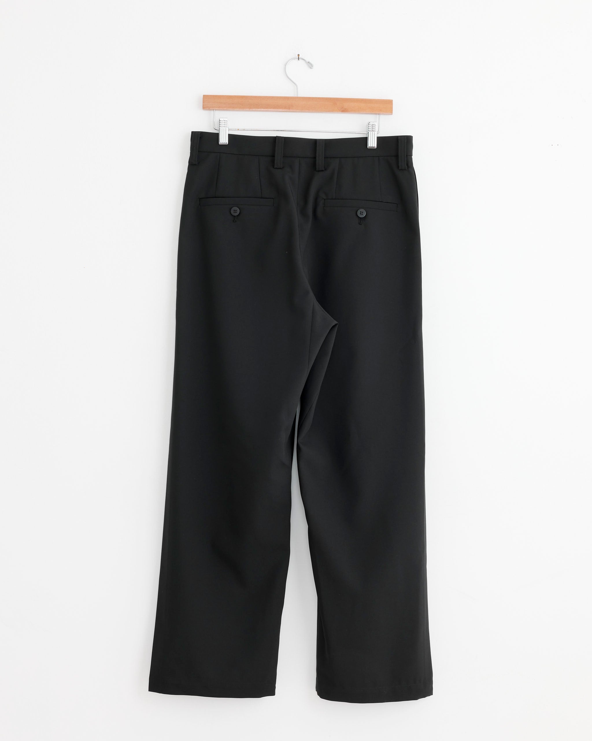 The IM MEN Wings Trouser in Black hangs on a wooden hanger against a white wall, shown from the back with two buttoned pockets, belt loops, and wrinkle-resistant polyester fabric visible.