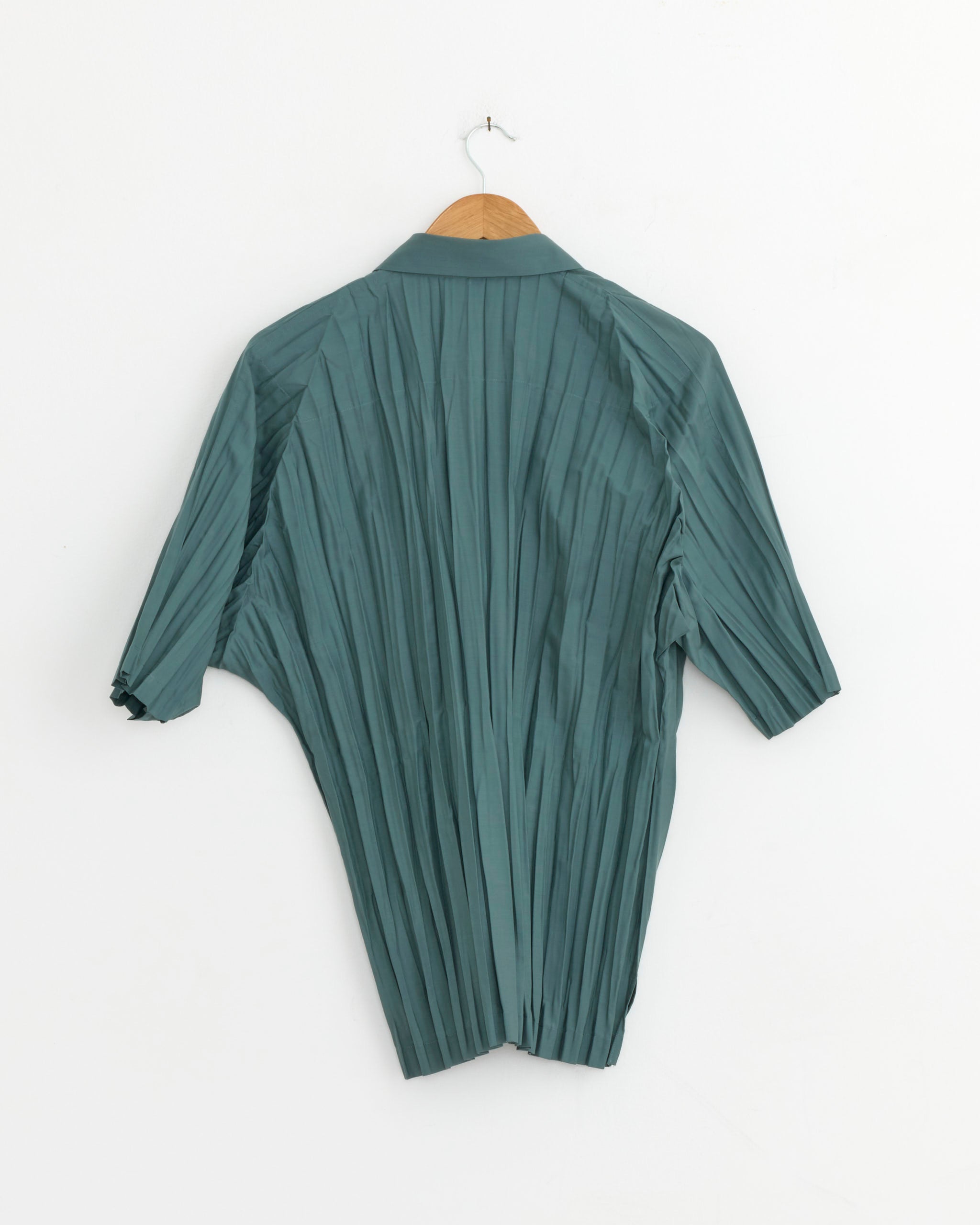 The IM MEN Wrinkle Shirt in Blue Grey, made in Japan from recycled polyester, hangs on a wooden hanger against a white wall with the back facing forward.