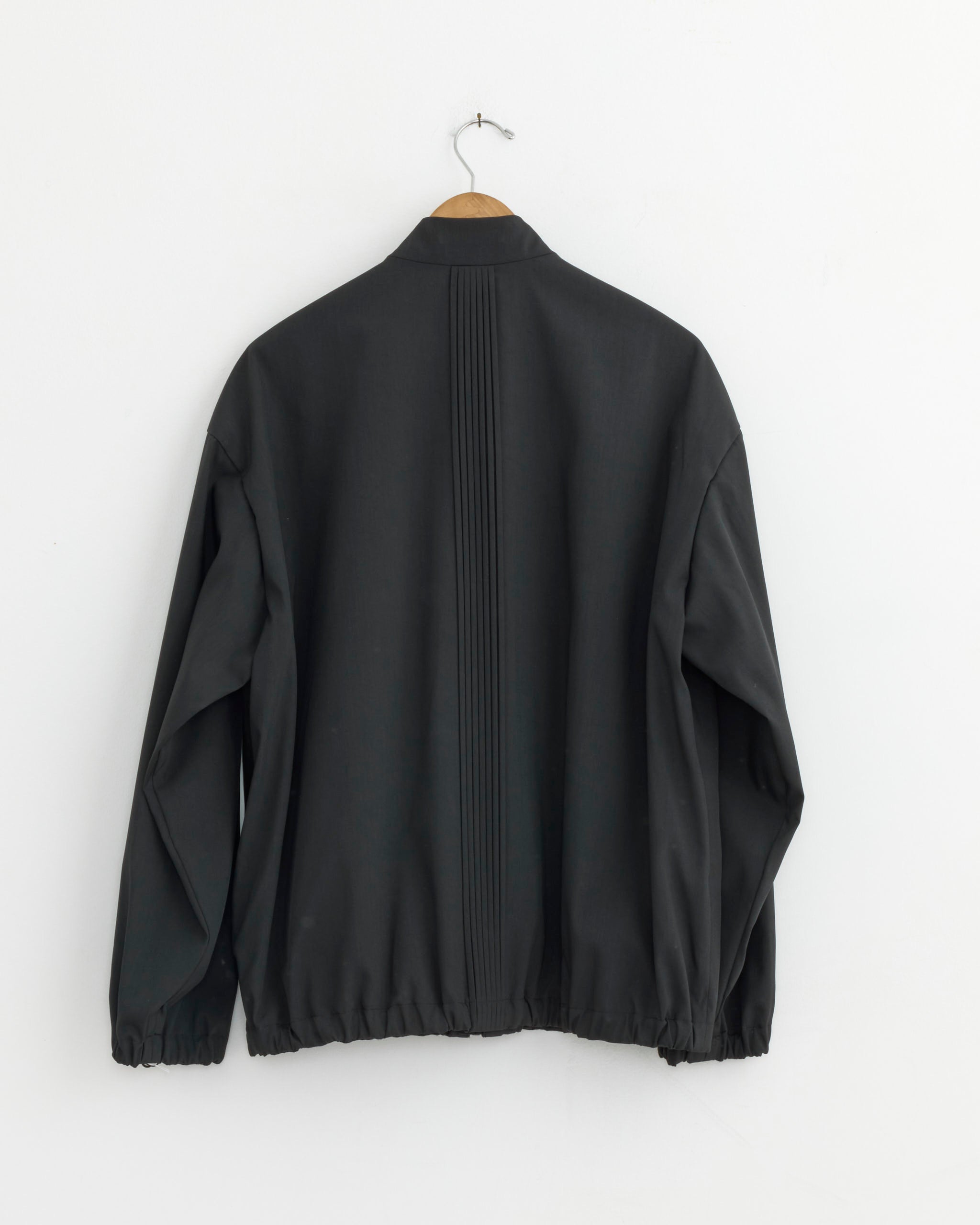 The IM MEN Air Pleats Jacket in Black hangs on a wooden hanger against a white wall, showing its lightweight, wrinkle-resistant design with a straight vertical pleat down the back.