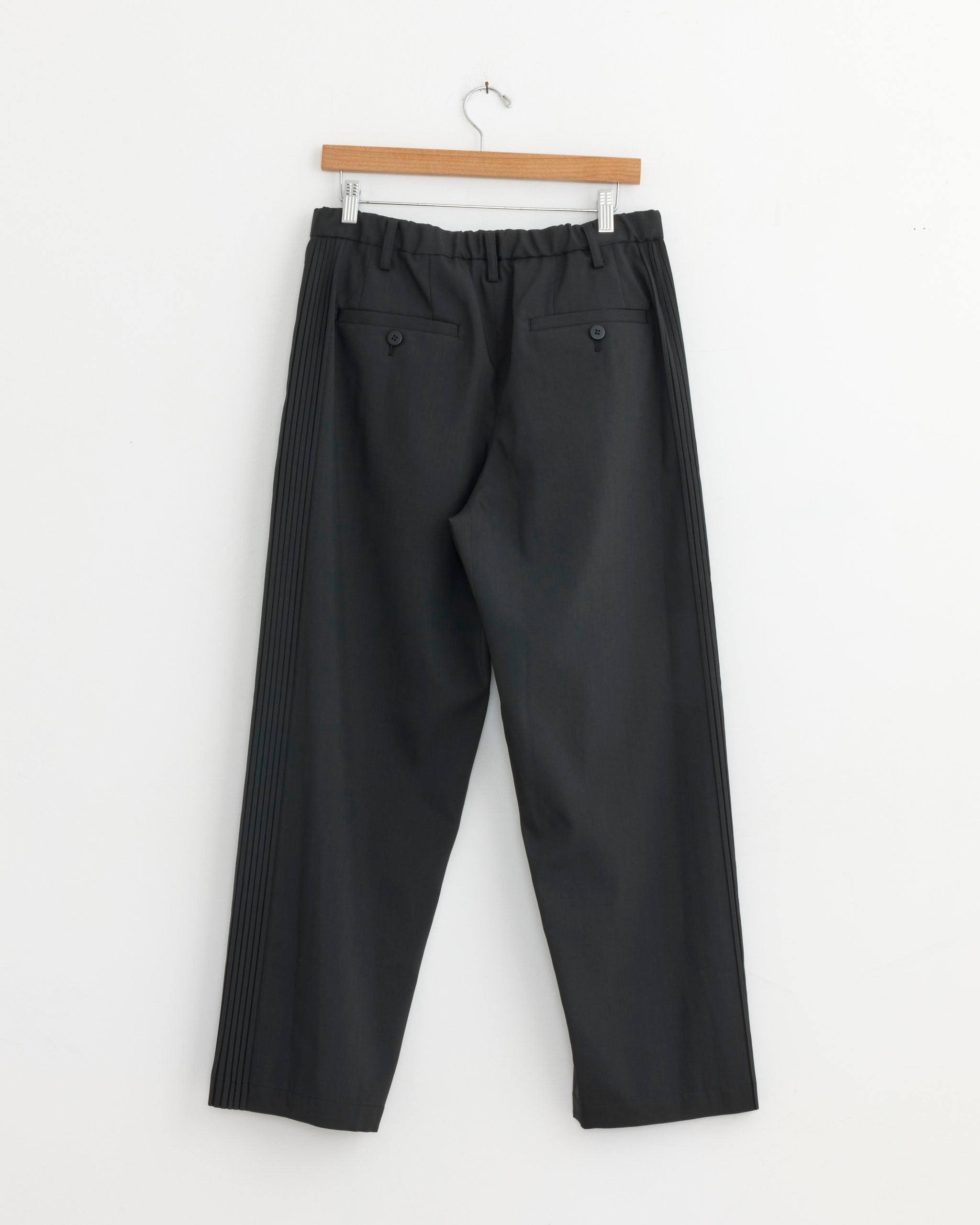 A pair of IM MEN Air Pleats Trouser in Black with an elastic waistband and buttoned back pockets, displayed on a wooden hanger against a white wall.