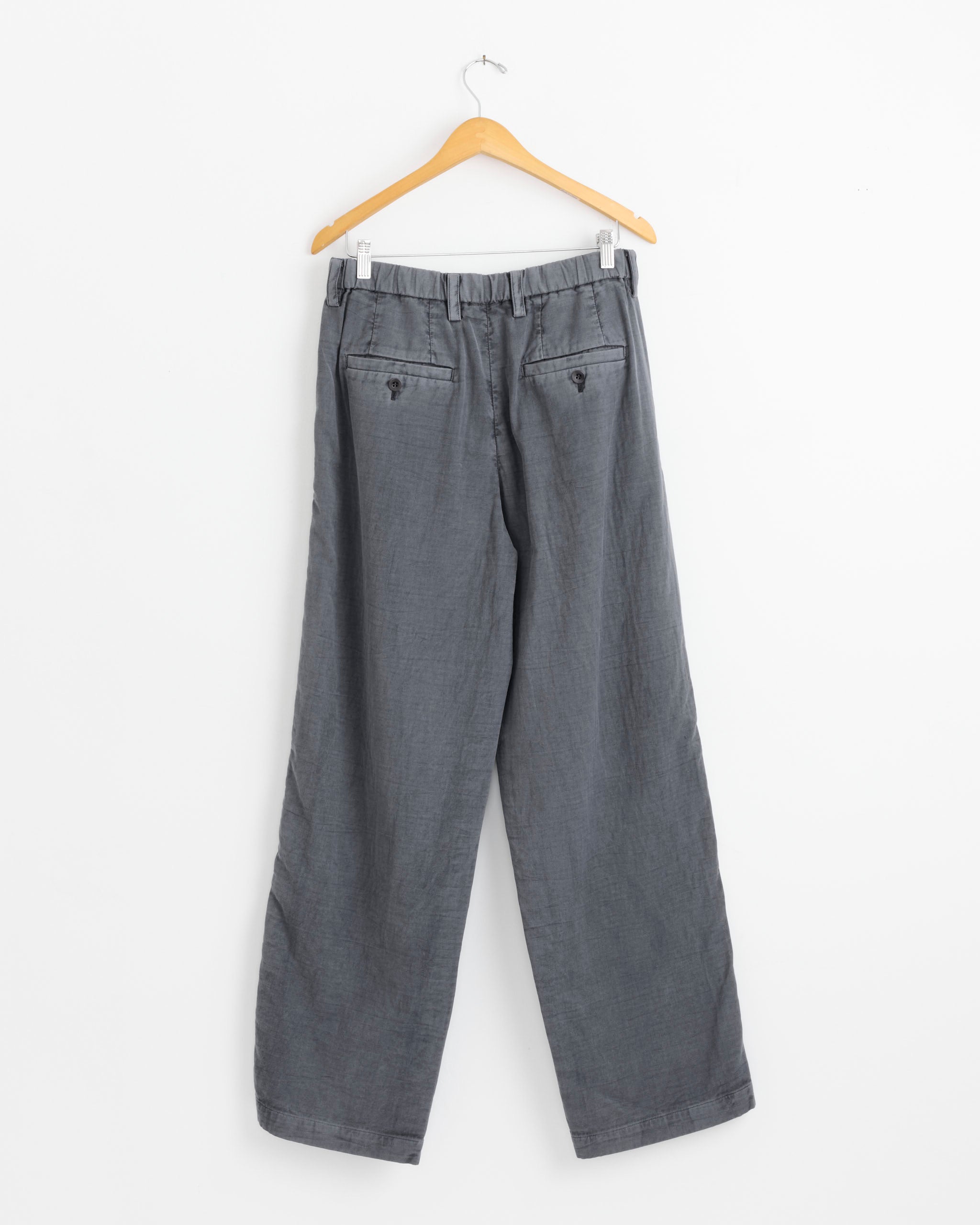 The IM MEN Pigment Double Face Pant in Charcoal Grey hangs on a wooden hanger before a white background, displayed from the back to show two buttoned pockets and belt loops.