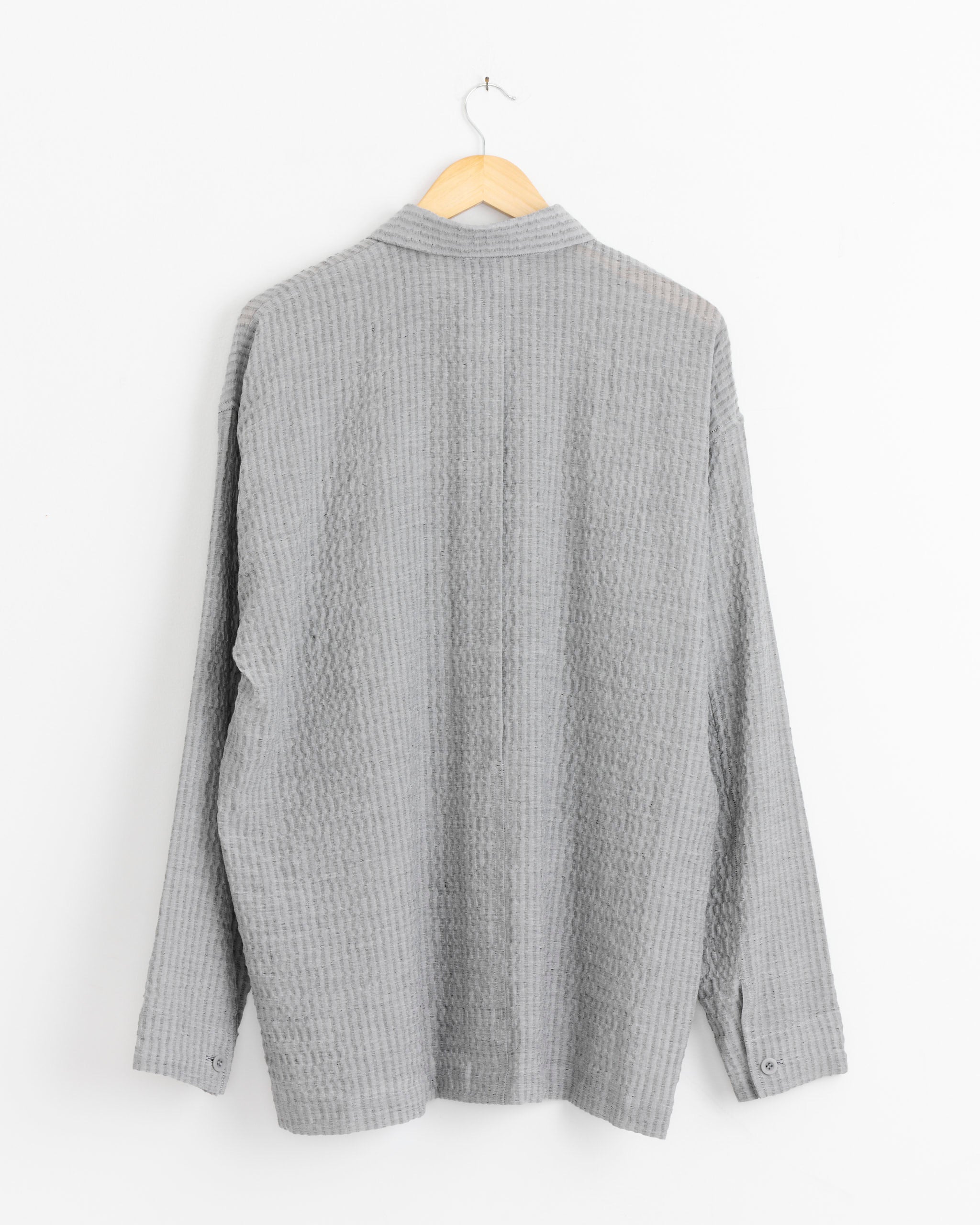 The IM MEN Stripe Shrink Shirt in Grey, featuring long sleeves and a textured finish, hangs on a wooden hanger against a white background with the back visible.