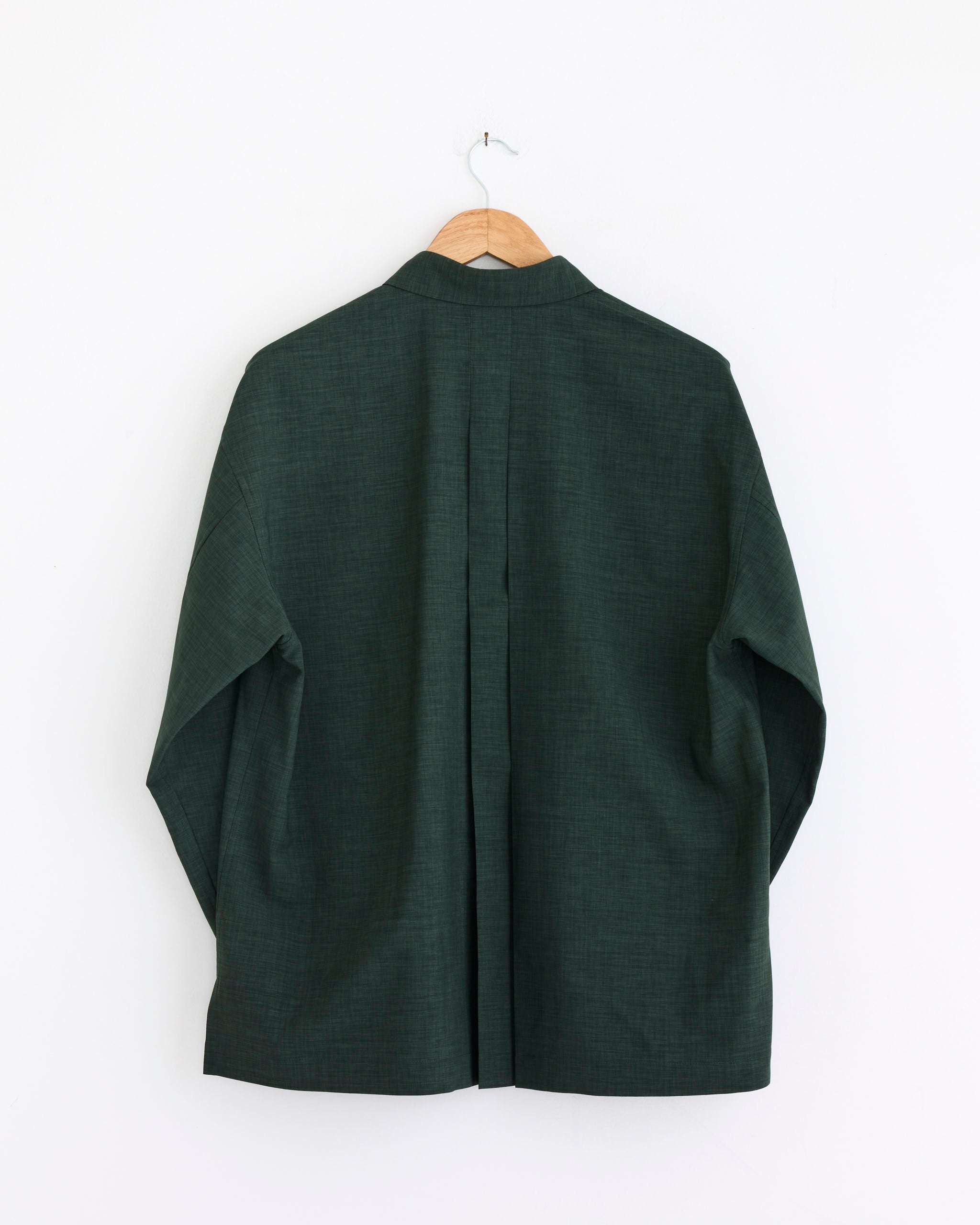 The IM MEN Linen Like Shirt in Dark Green, shown from the back, hangs on a wooden hanger against a white wall. This long-sleeve shirt features a loose fit and center pleat.