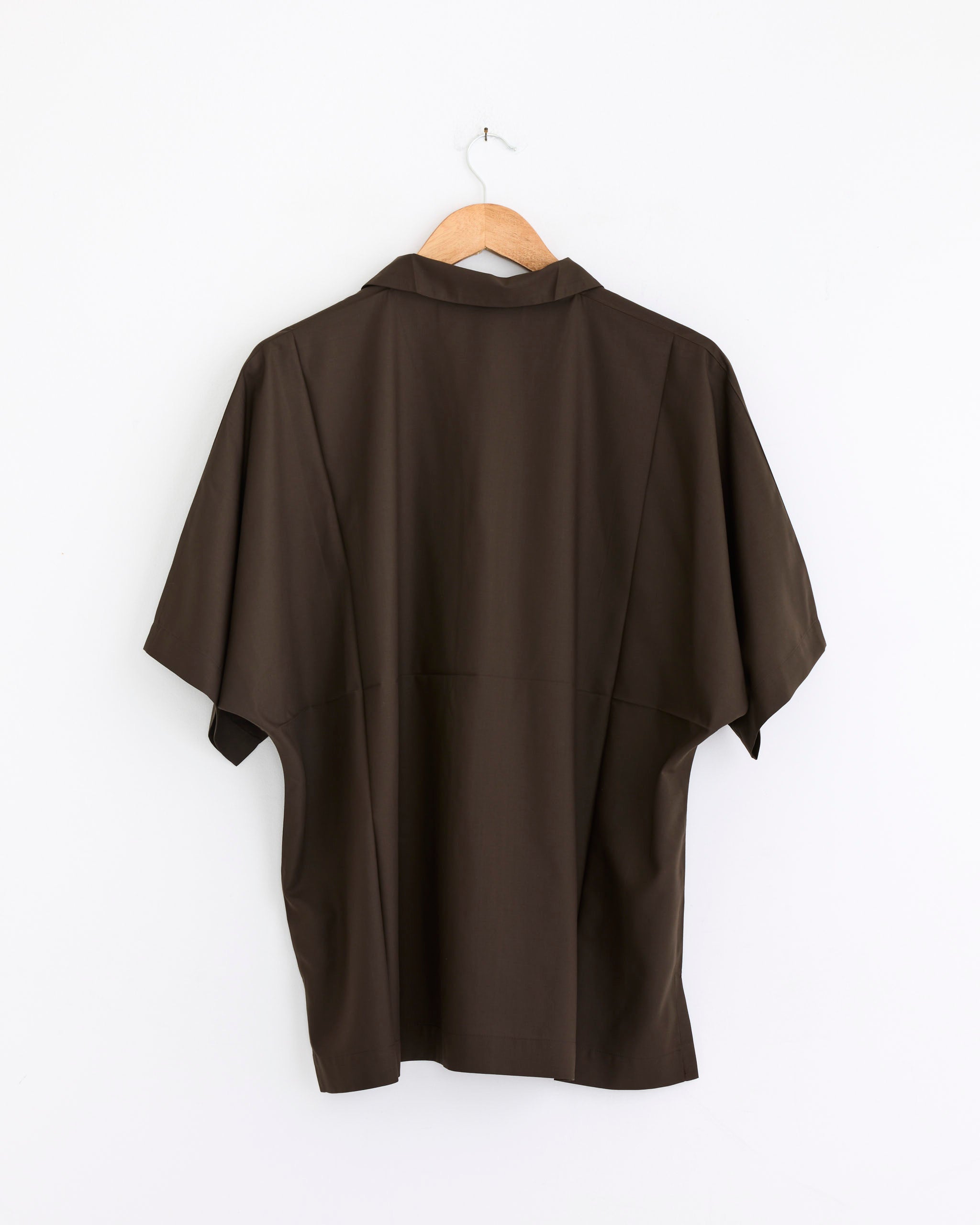 A wrinkle-resistant, short-sleeved Compact Shirt in dark brown by IM MEN hangs on a wooden hanger against a plain white wall, shown from the back.