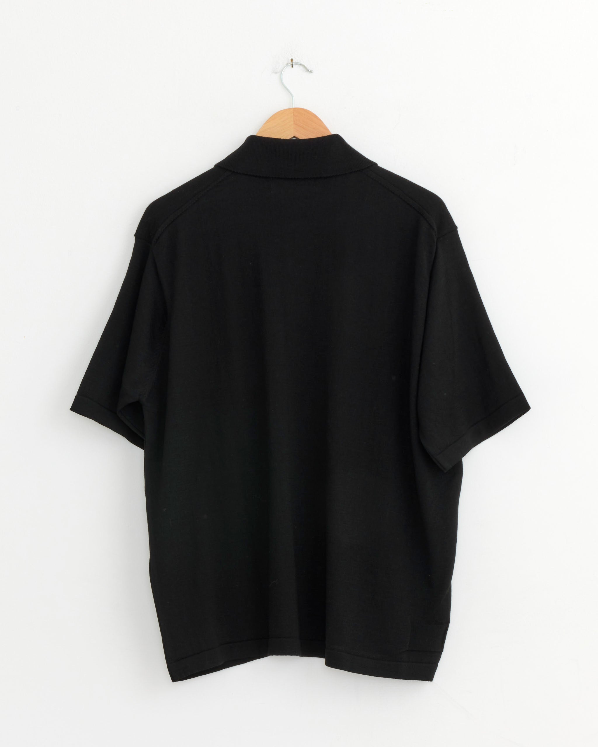 The Universal Works Knit Shirt in Black is displayed from the back, hanging on a wooden hanger against a white wall.