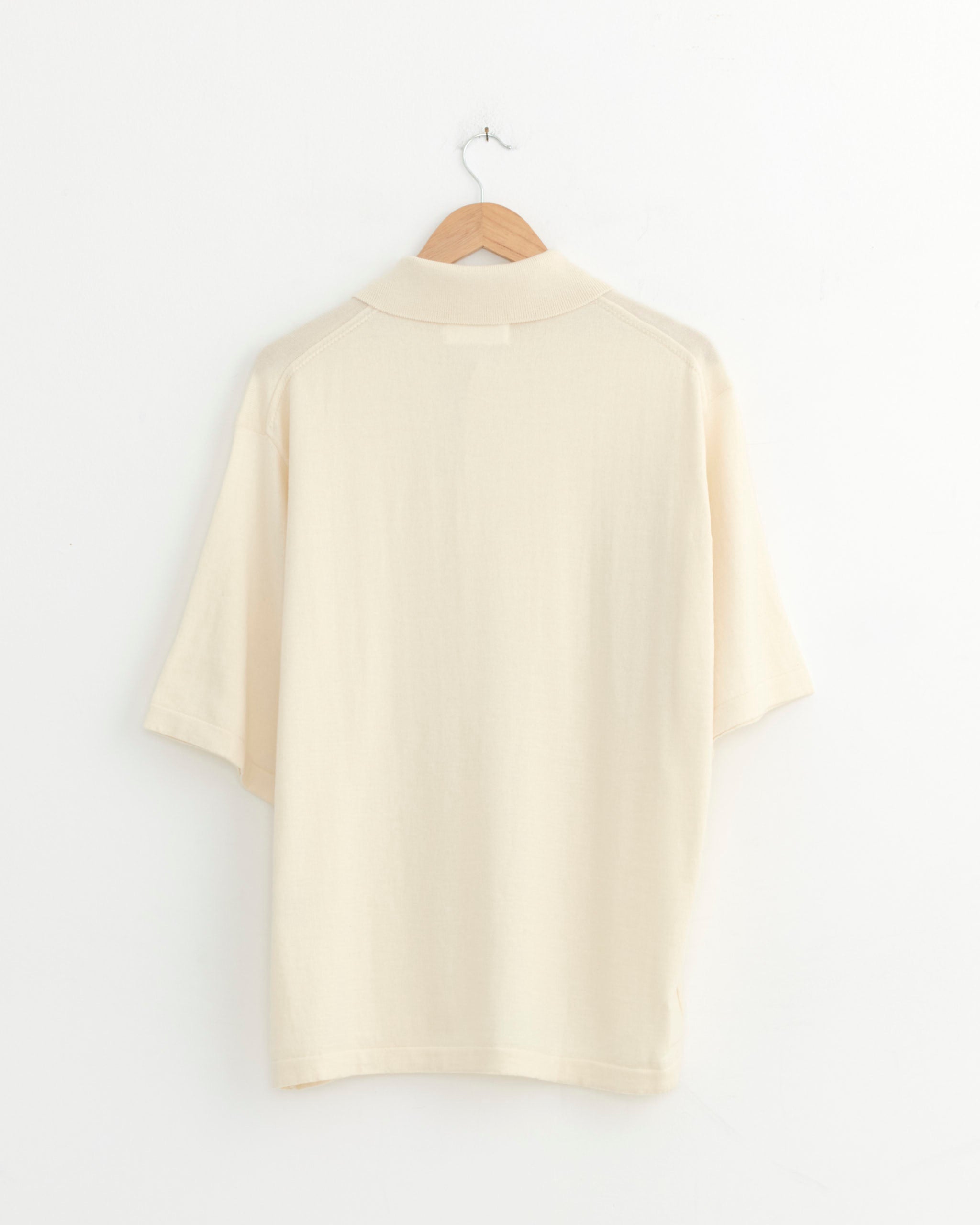 The Universal Works Knit Shirt in Ecru, a short-sleeve organic cotton knit, hangs on a wooden hanger against a plain white wall, displayed from the back.