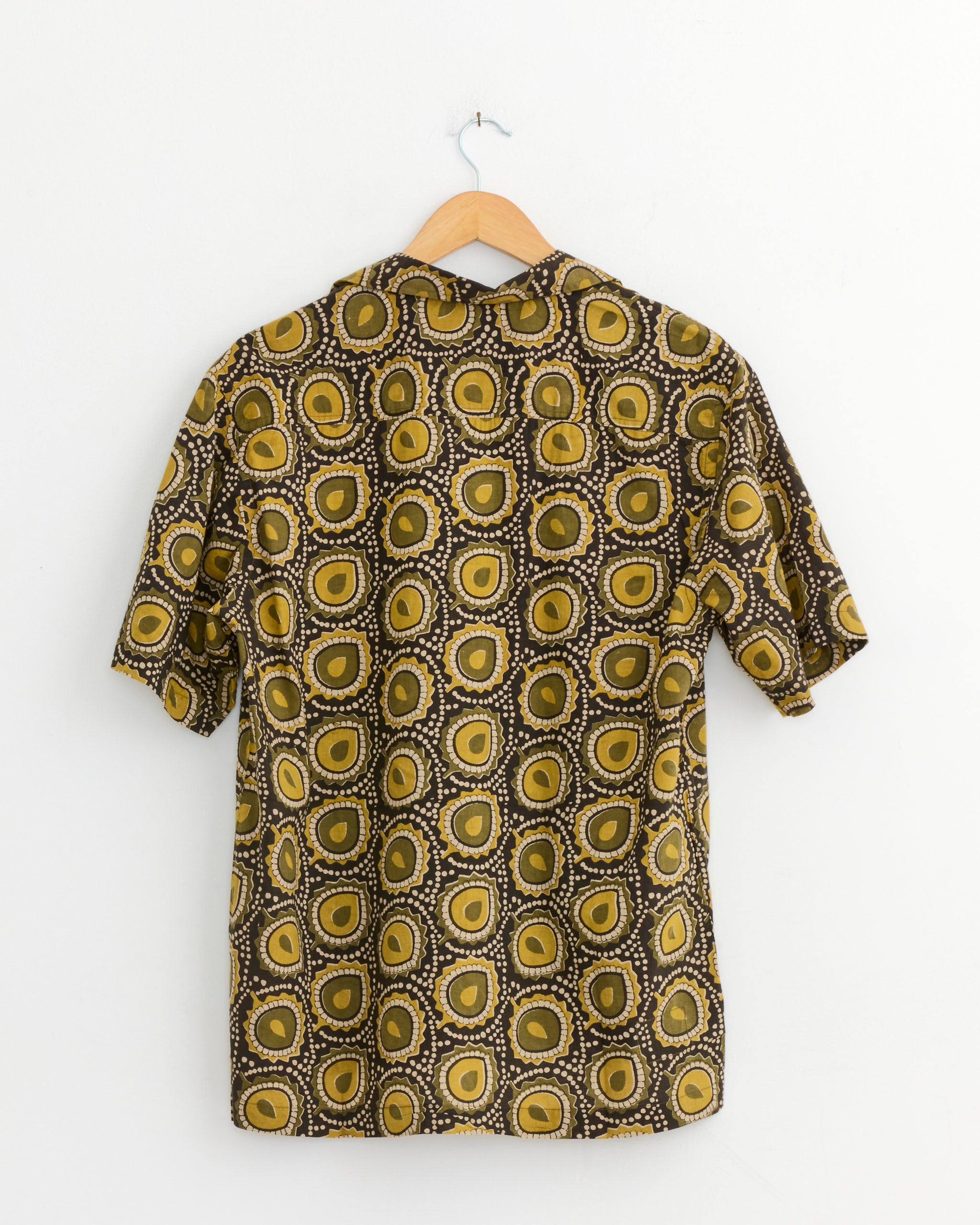 The Universal Works Camp Shirt in Olive, featuring a lightweight cotton fabric and an olive circular pattern with yellow, brown, and black accents, hangs on a wooden hanger against a white wall.