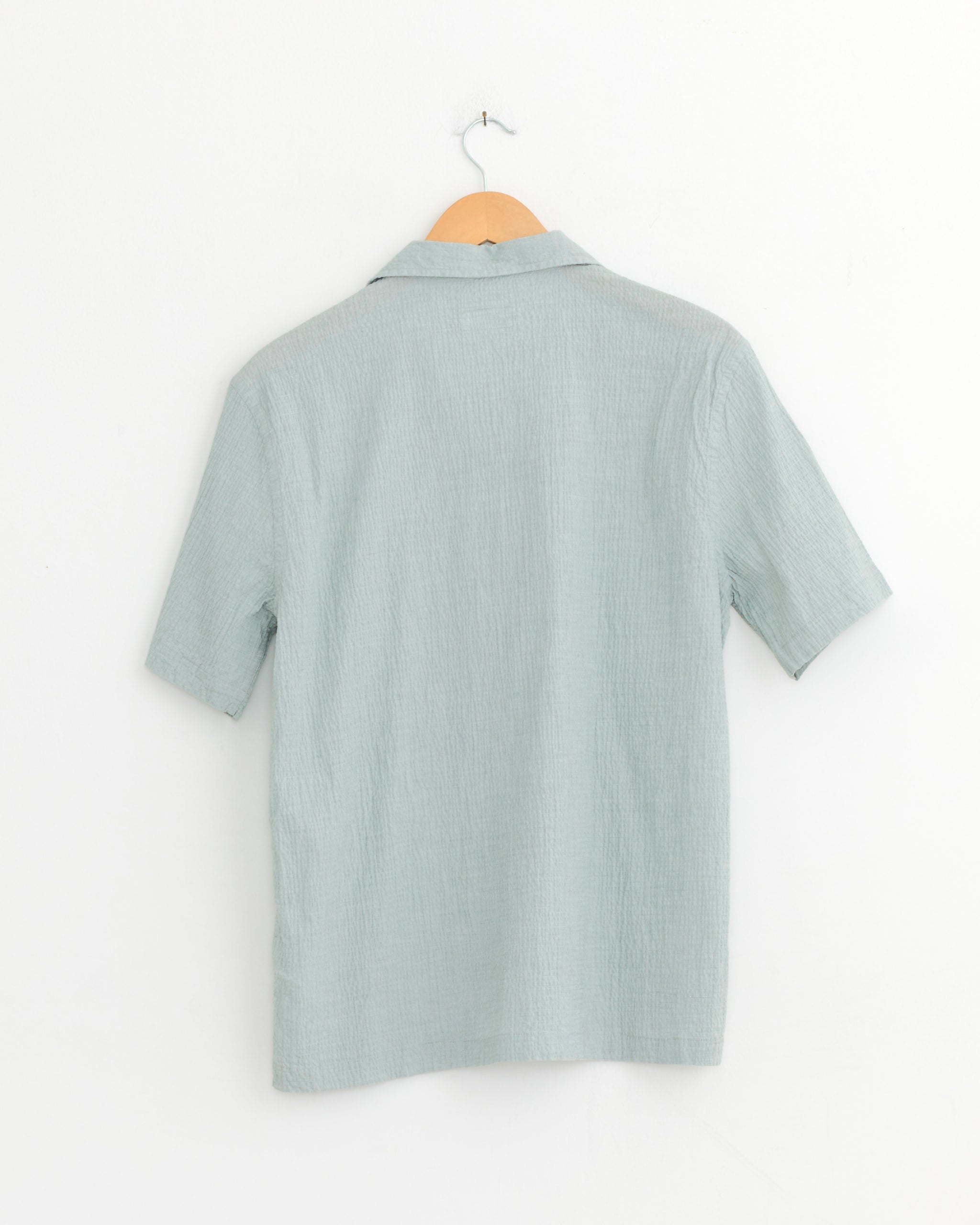A Universal Works Road Shirt in green with short sleeves hangs on a wooden hanger against a plain white wall, shown from the back.