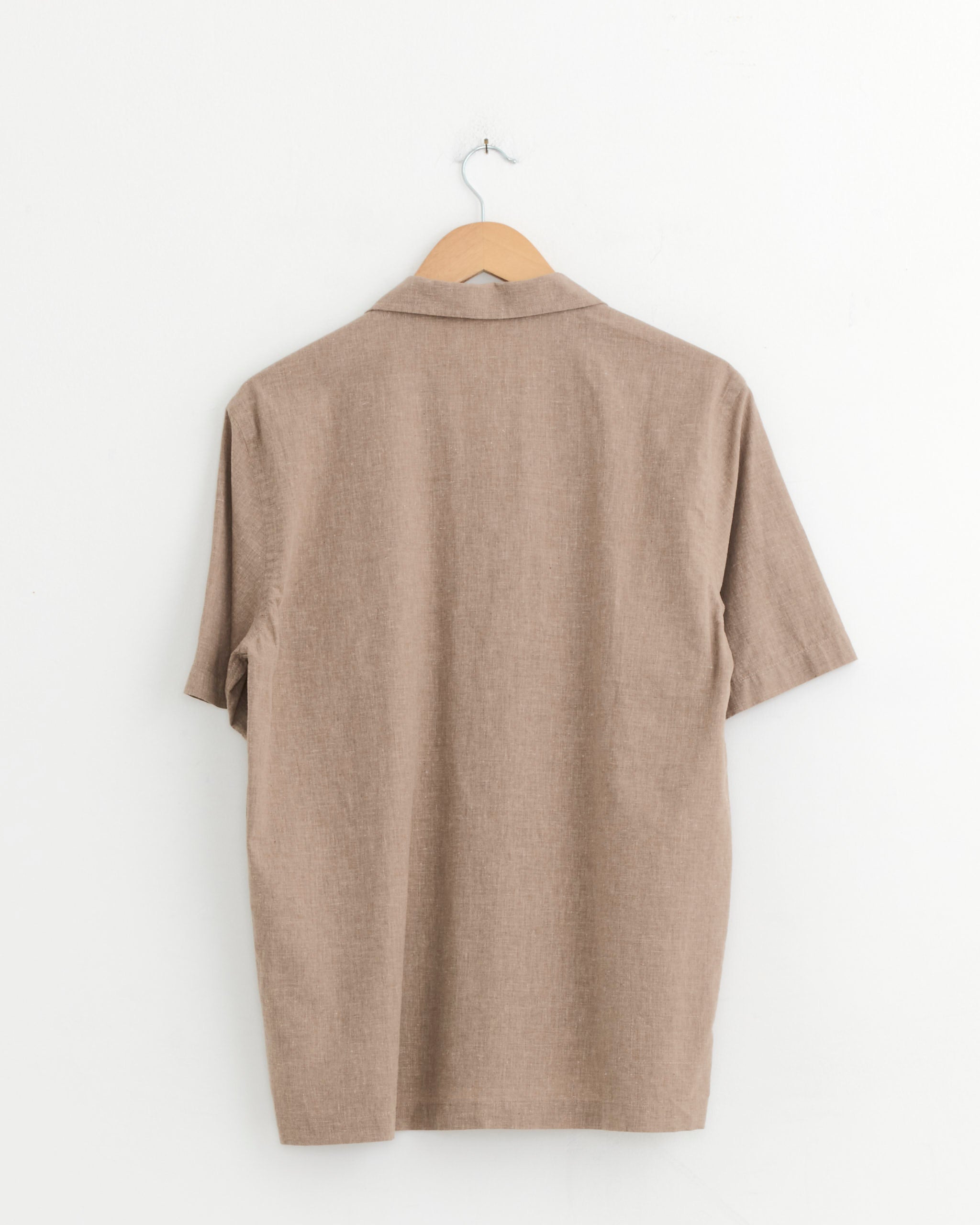 The Universal Works Road Shirt in Brown, made from cotton linen with short sleeves, is displayed from the back hanging on a wooden hanger against a plain white wall.