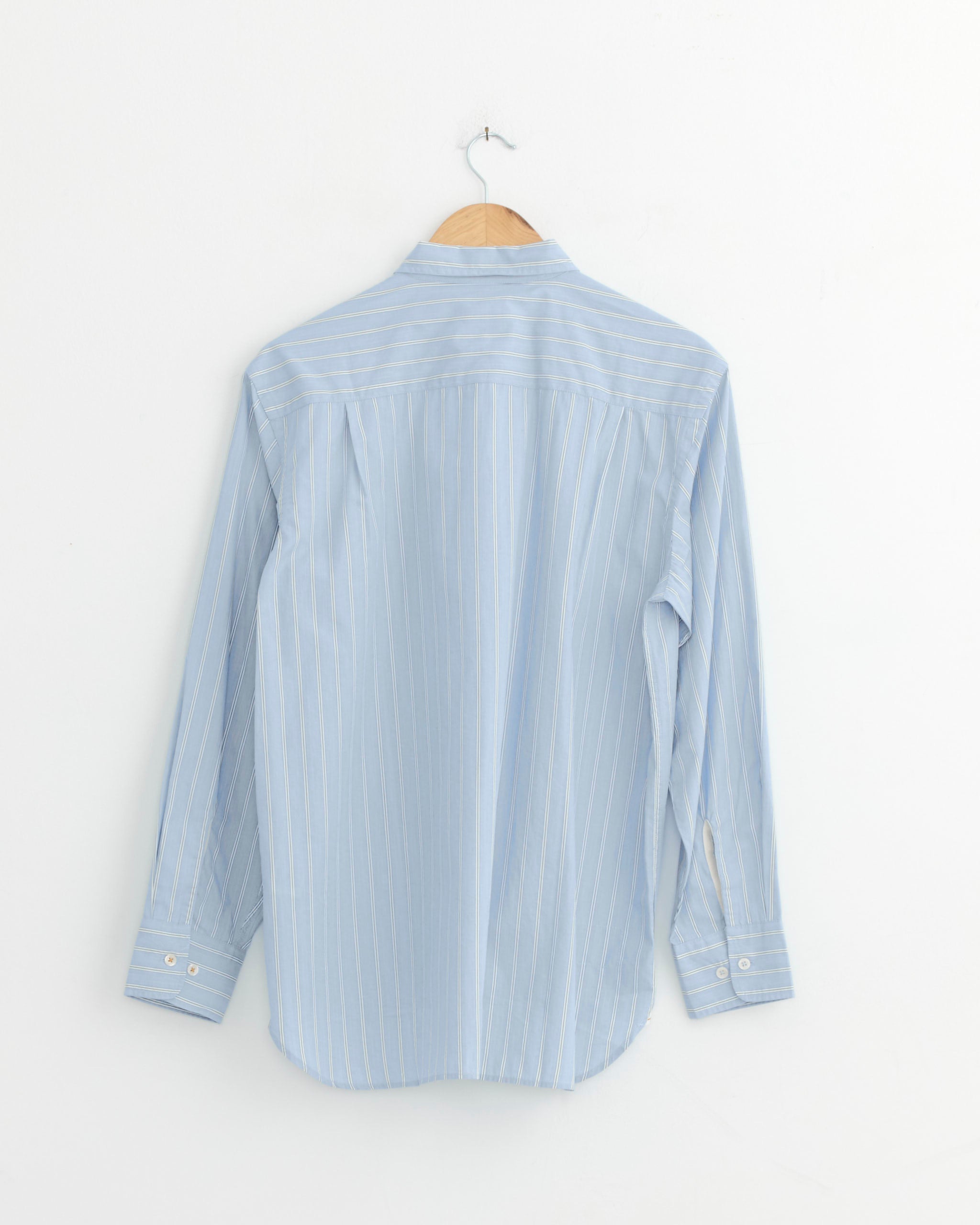 A Universal Works Square Pocket Shirt in Blue, featuring long sleeves, 100% cotton fabric, and white vertical stripes, hangs on a wooden hanger against a white wall with the back side visible.