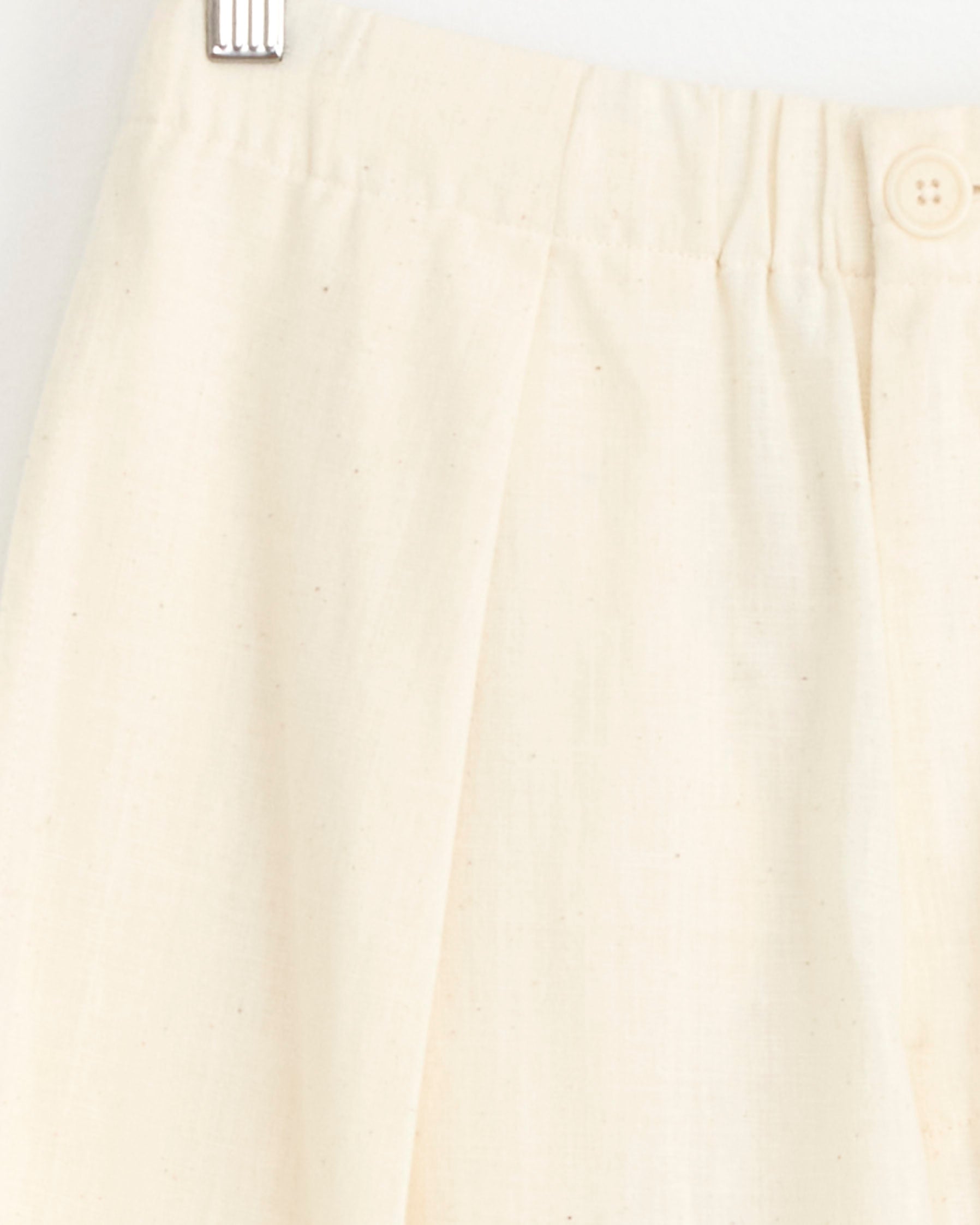 Close-up of the Kardo Taka Trouser in Natural, featuring cream-colored corduroy with an elastic waistband, single button, and subtle pleats, displayed on a white hanger.