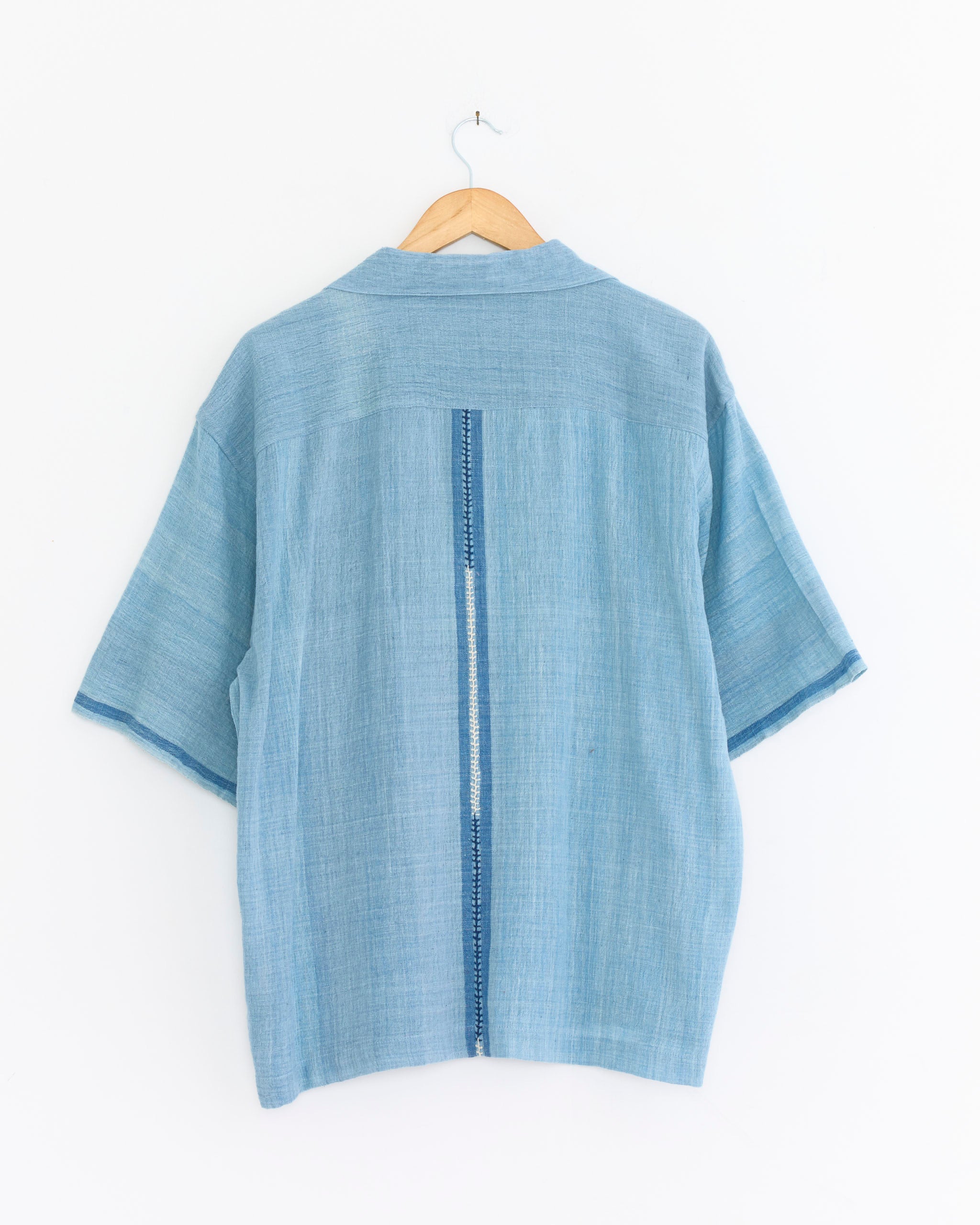 The Rohit Shirt in Light Blue by Kardo is a handloom cotton shirt with short sleeves, a straight hem, and a vertical stitched detail down the back, shown on a wooden hanger against a white background.