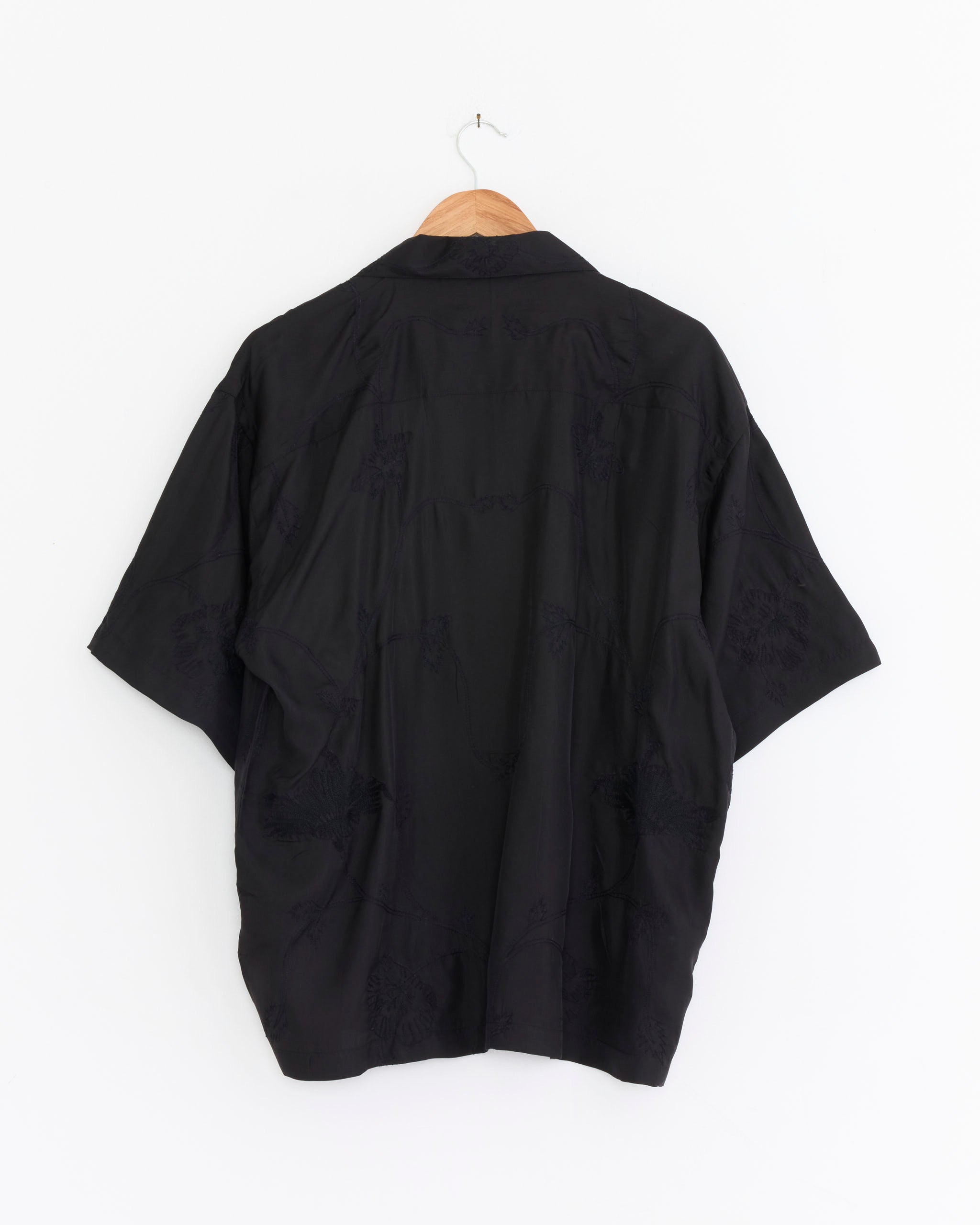 The Rohit Shirt in Black by Kardo, crafted from silk with a camp collar and made in India, hangs on a wooden hanger against a white background, displayed from the back.