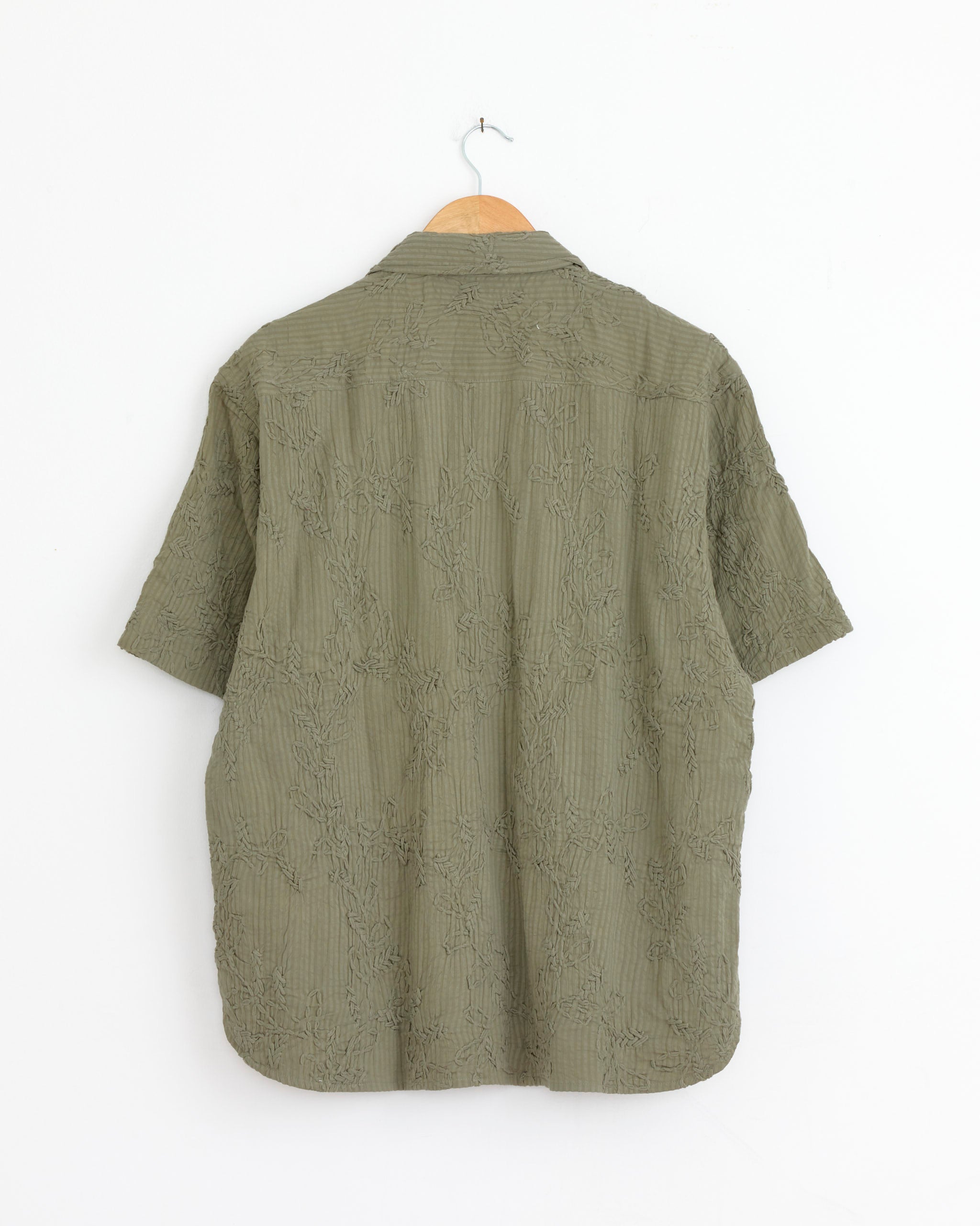 The Kardo Ronen Shirt in Olive, a khadi cotton short-sleeve button-up with textured embroidery and a relaxed camp collar, hangs on a wooden hanger against a plain white wall, shown from the back.