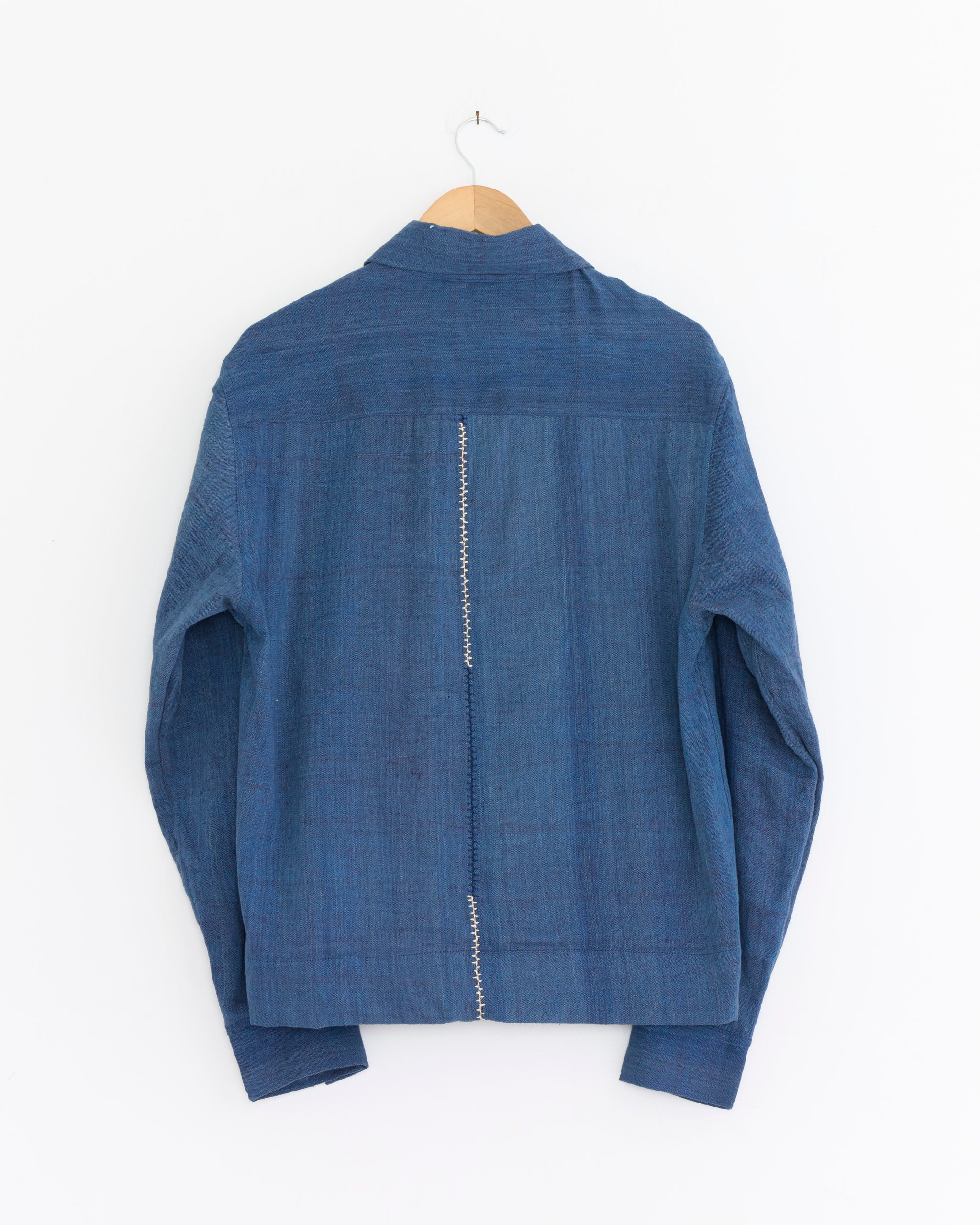 The Bodhi Jacket in Blue by Kardo, crafted from handloom cotton with a visible vertical seam down the back, hangs on a wooden hanger against a plain white wall.