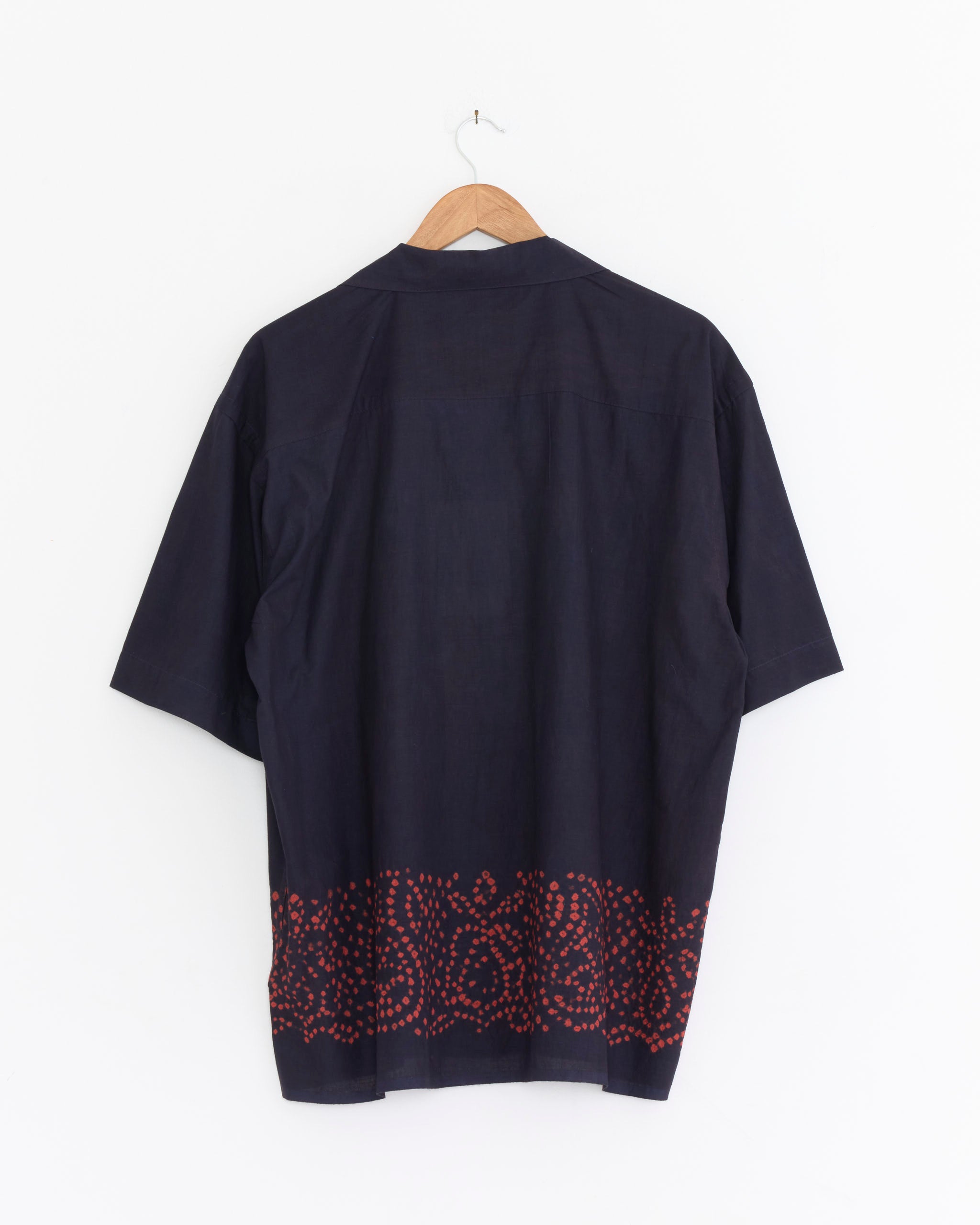 The Kardo Rohit Shirt in Oxblood, made from naturally dyed cotton, features short sleeves, a camp collar, and a red geometric pattern along the bottom hem. It hangs on a wooden hanger against a white background.