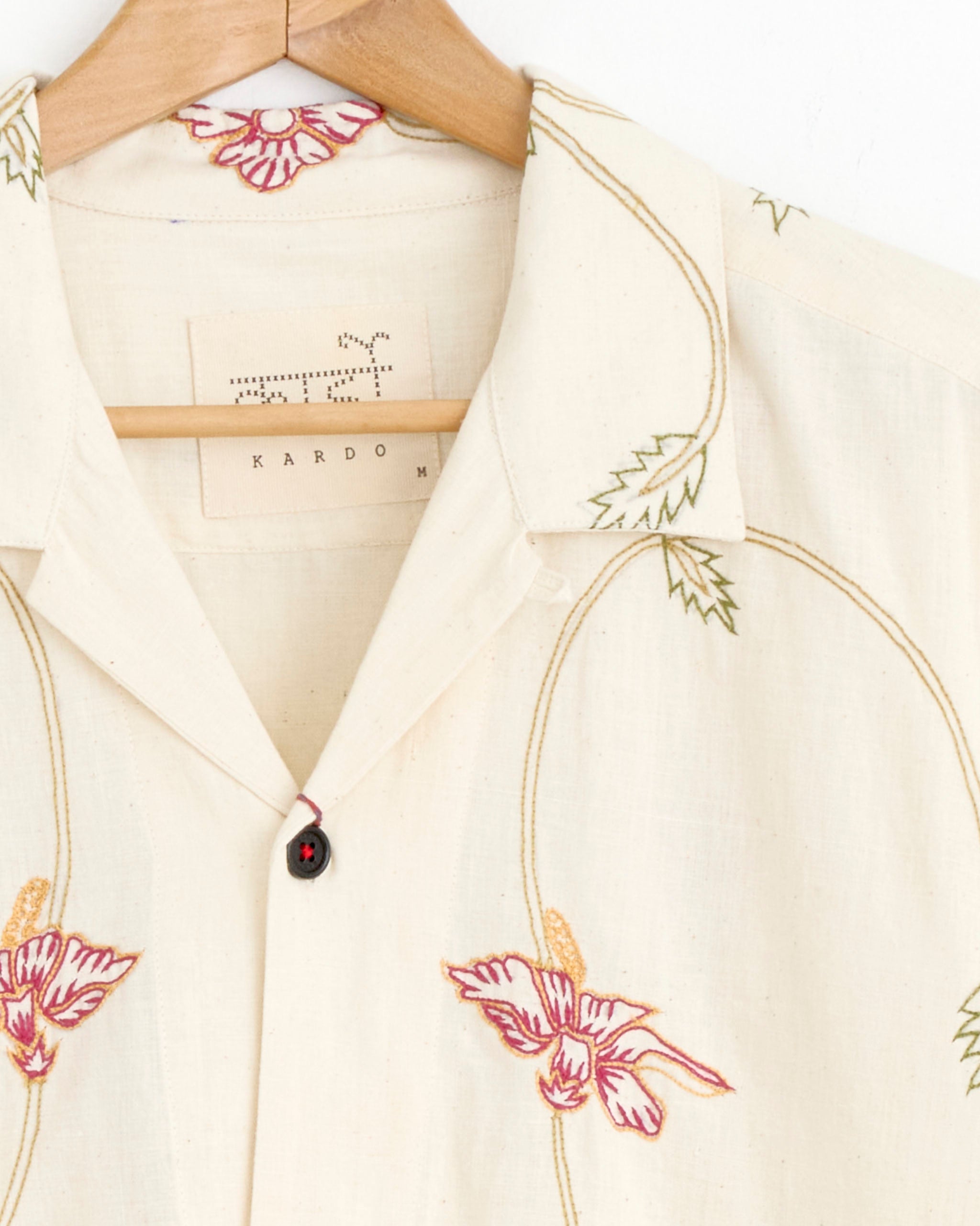 The Ronen Shirt in Natural by Kardo is a cream khadi cotton collared shirt with red, pink, yellow, and green floral embroidery, shown on a wooden hanger. The inner label displays the brand name Kardo.