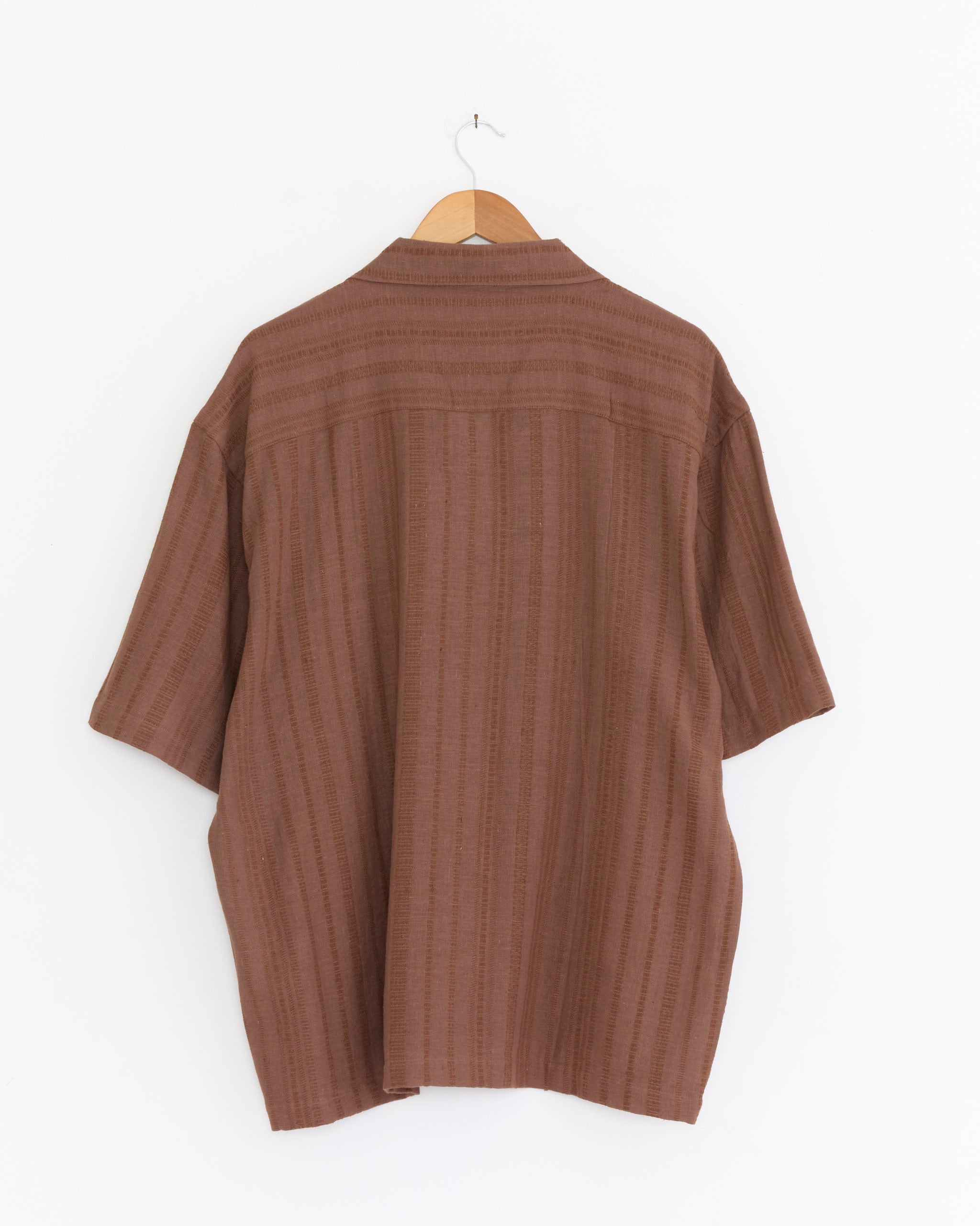 The Kardo Rohit Shirt in Ganache, featuring short sleeves, a camp collar, and subtle stripes, is displayed from the back on a wooden hanger against a plain white background.