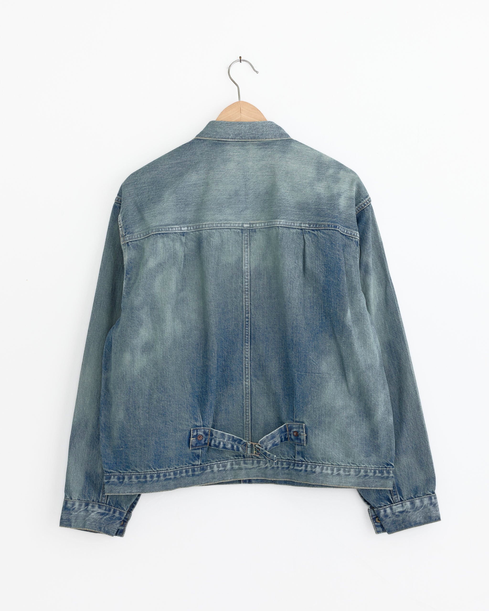 An A.PRESSE 1st Type Denim Jacket in Indigo hangs on a wooden hanger, displayed from the back against a white wall. The jacket showcases visible seams, vintage fading on the shoulders and sleeves, and timeless classic styling.