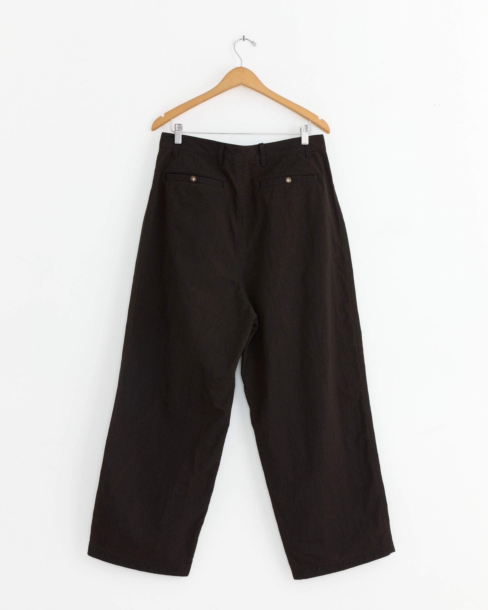 The 003 Pant in Brown by Comme Des Garçons Homme hangs on a wooden hanger against a white wall.