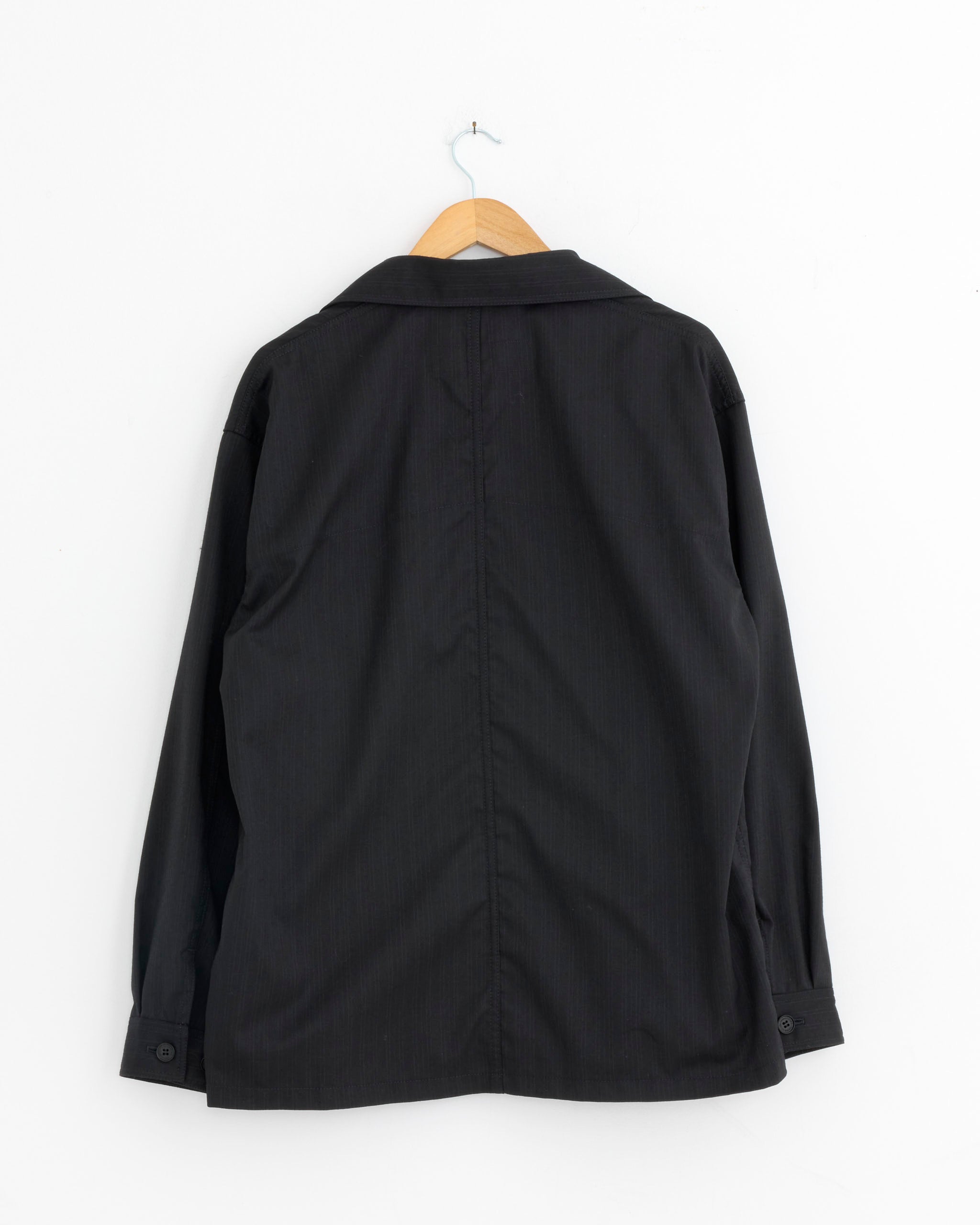 The 005 Jacket in Navy by Comme Des Garçons Homme hangs on a wooden hanger against a white wall, displayed from the back with a visible center seam.