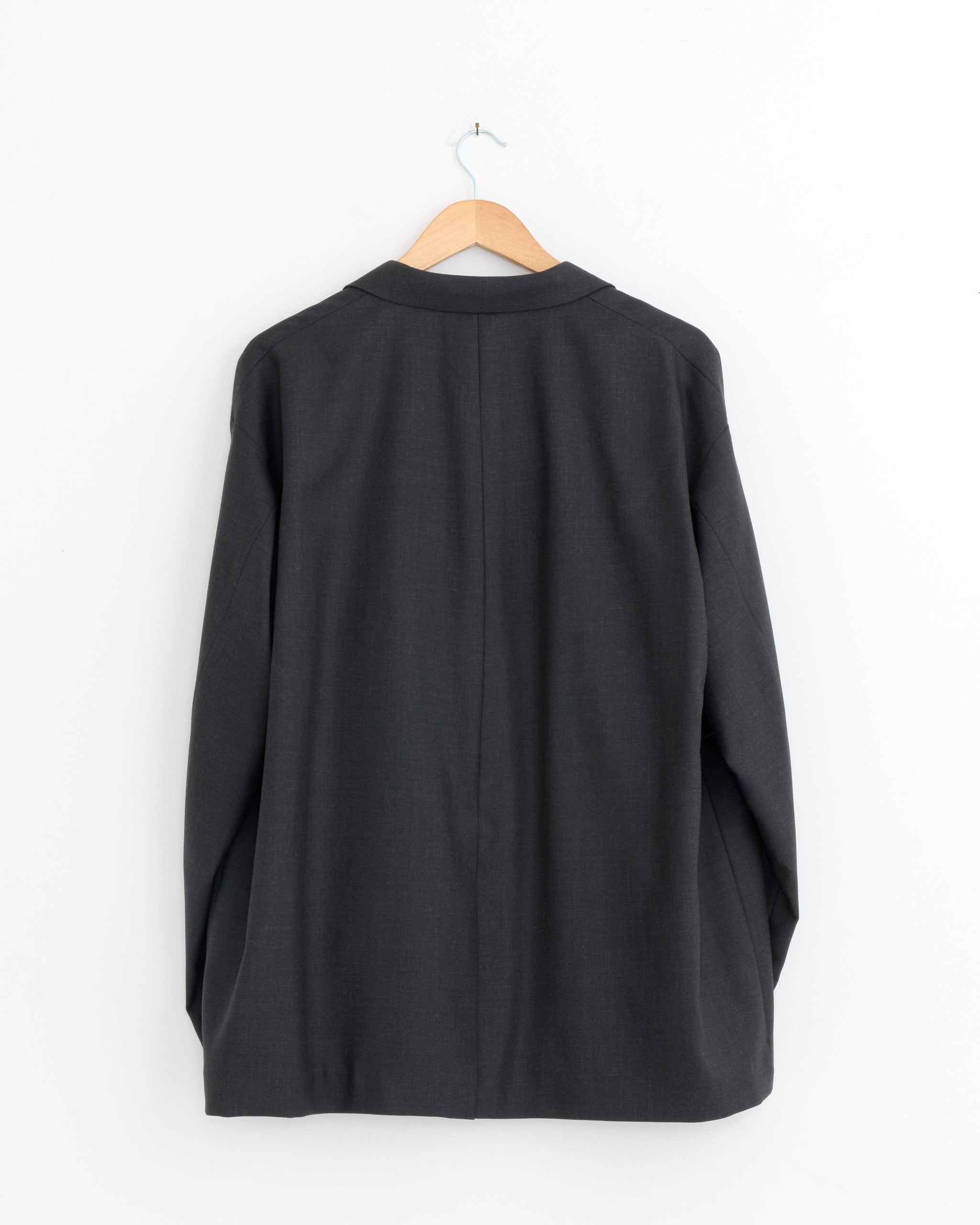 The Comme Des Garçons Homme 009 Jacket in Charcoal, 100% wool, size Large X-Large, is displayed from the back on a wooden hanger against a white wall.