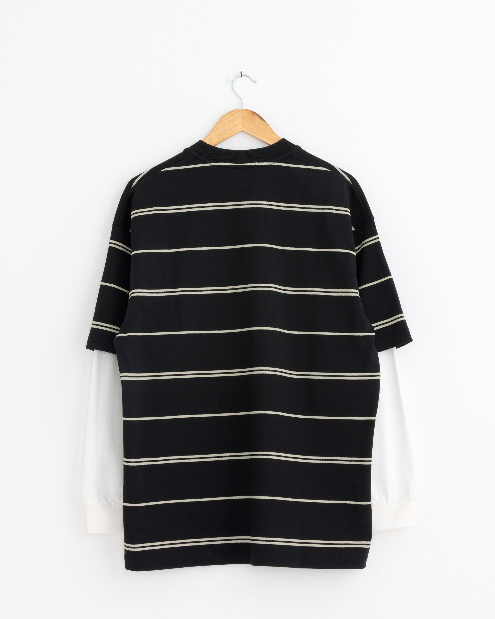 The Comme Des Garçons Homme 003 T-Shirt in Black/Grey/Off White, made from 100% Japanese cotton, is layered over a white long-sleeve shirt and displayed on a wooden hanger against a white wall.