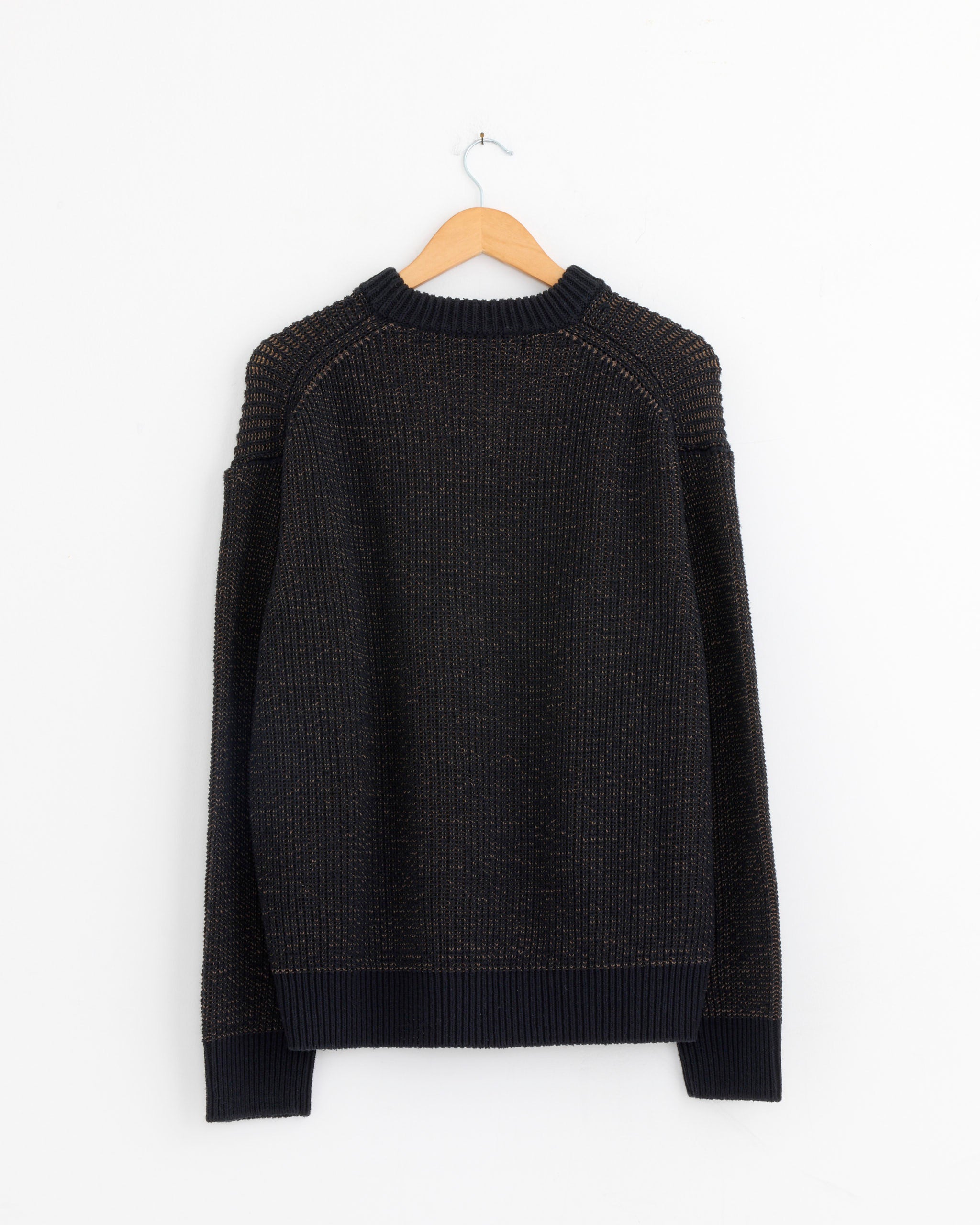 The Comme Des Garçons Homme 001 Sweater in Dark Navy/Brown hangs on a wooden hanger against a white wall. Made from Japanese fabric, it has a ribbed texture, long sleeves, and a classic crew neckline.
