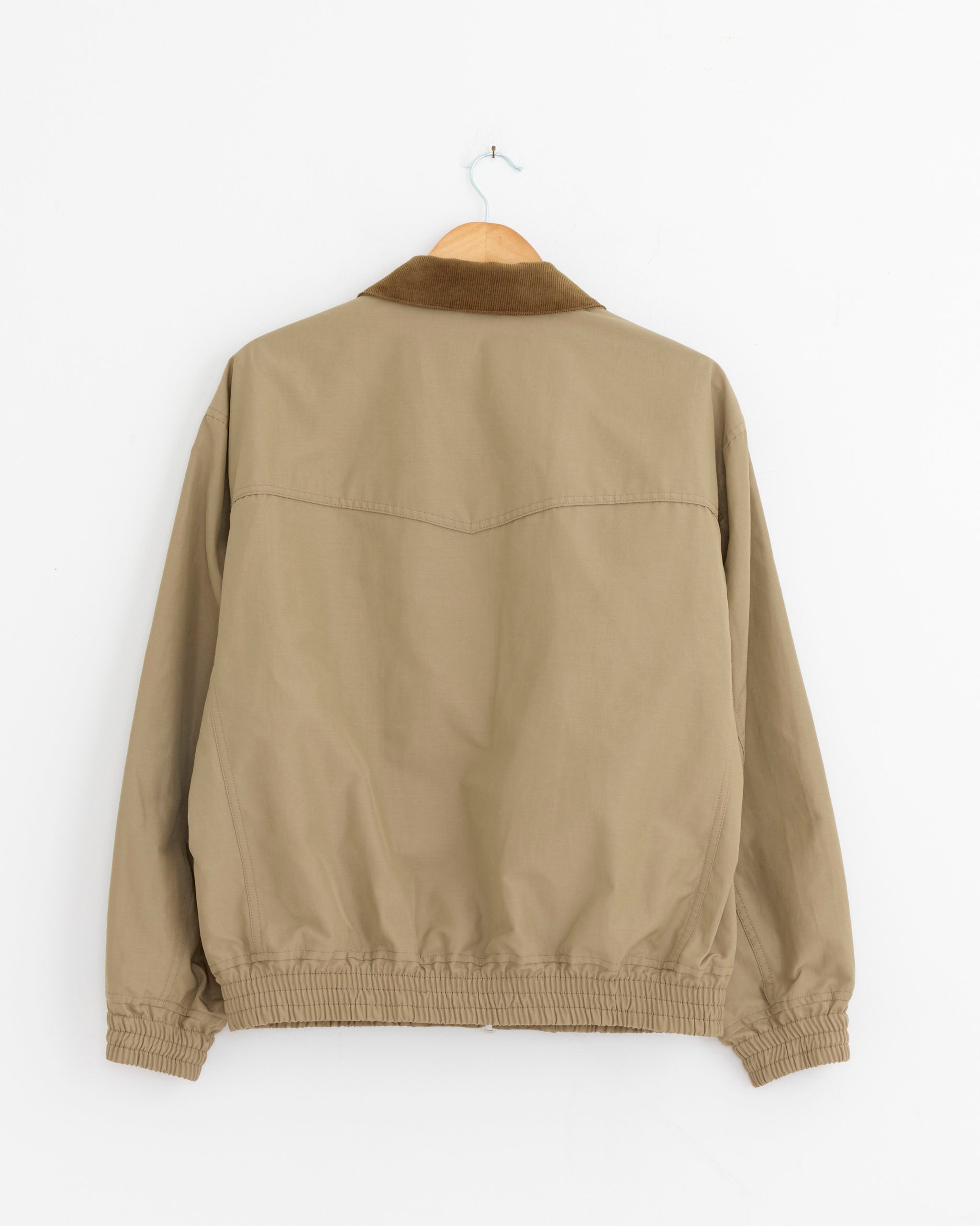 The Comme Des Garçons Homme 032 Jacket in Beige, made from polyester linen with a darker brown collar and elastic cuffs and hem, hangs on a wooden hanger against a white wall, shown from the back.