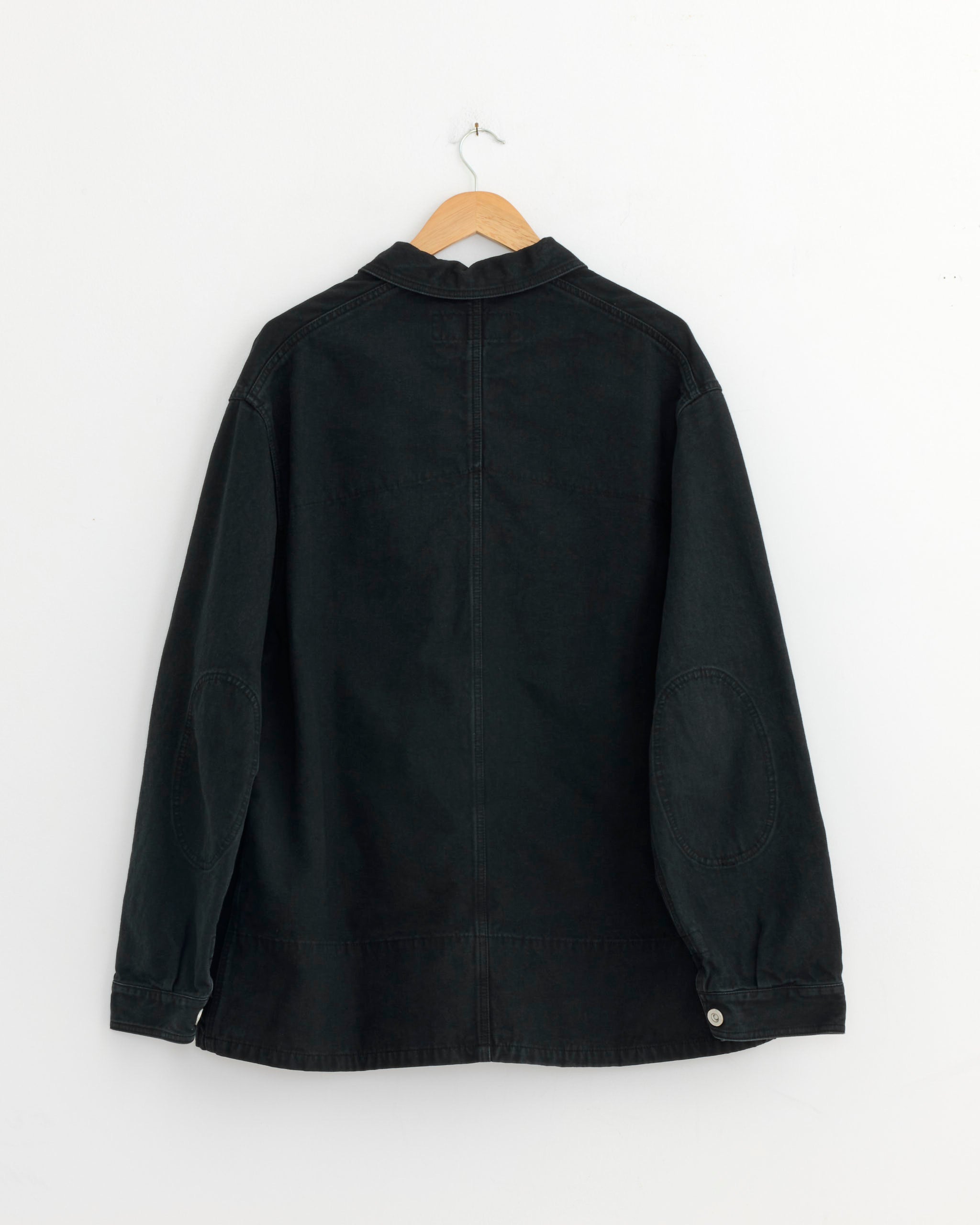 The Comme Des Garçons Homme 021 Jacket in Black Mix, crafted from cotton denim in Japan, hangs on a wooden hanger against a white wall. Shown from the back, it features visible stitching, a collar, and silver cuff buttons.