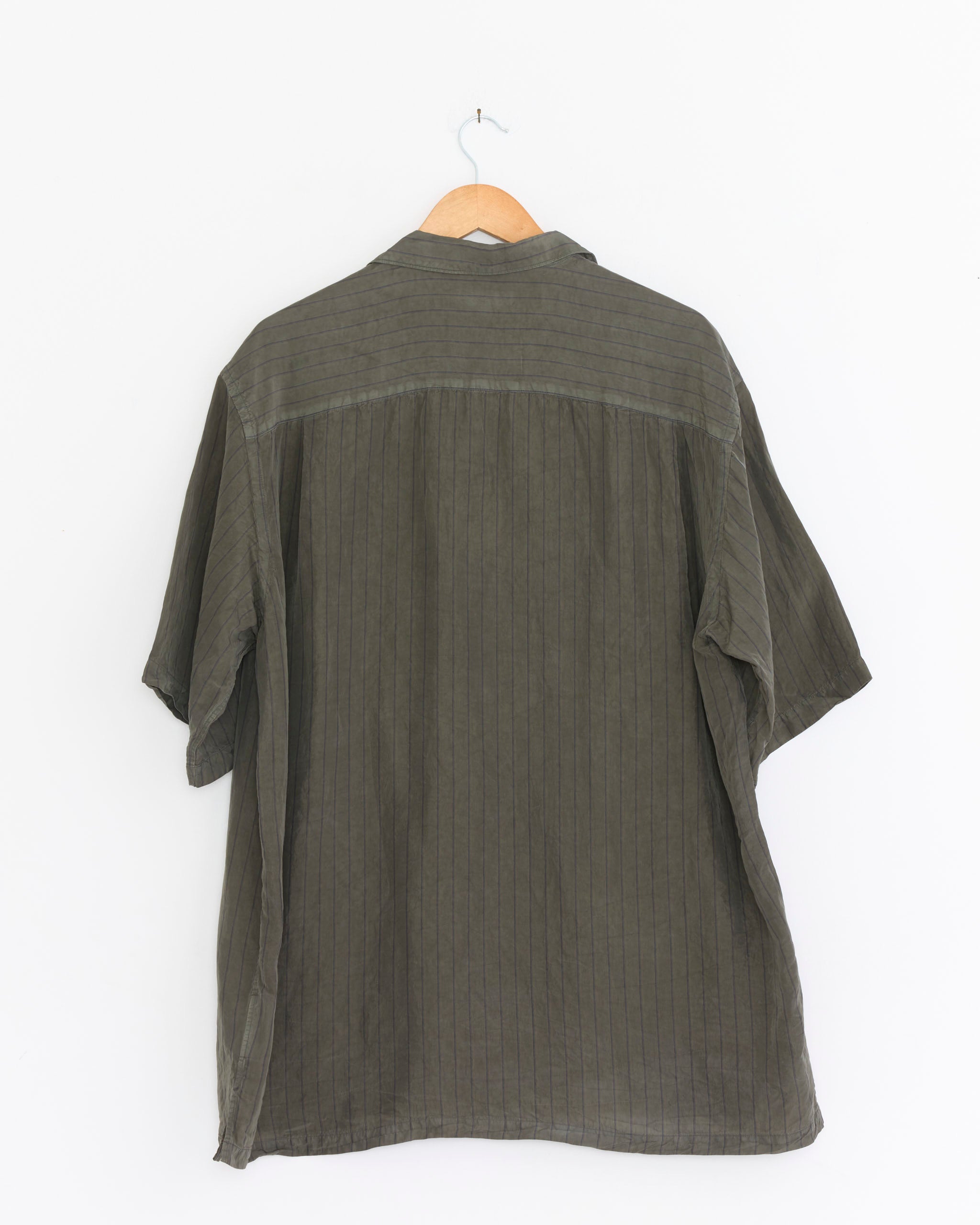 The Comme Des Garçons Homme 023 Shirt in Khaki, a loose-fitting, short-sleeved camp collar shirt with a subtle pinstripe pattern, is shown from the back on a wooden hanger against a plain white background.