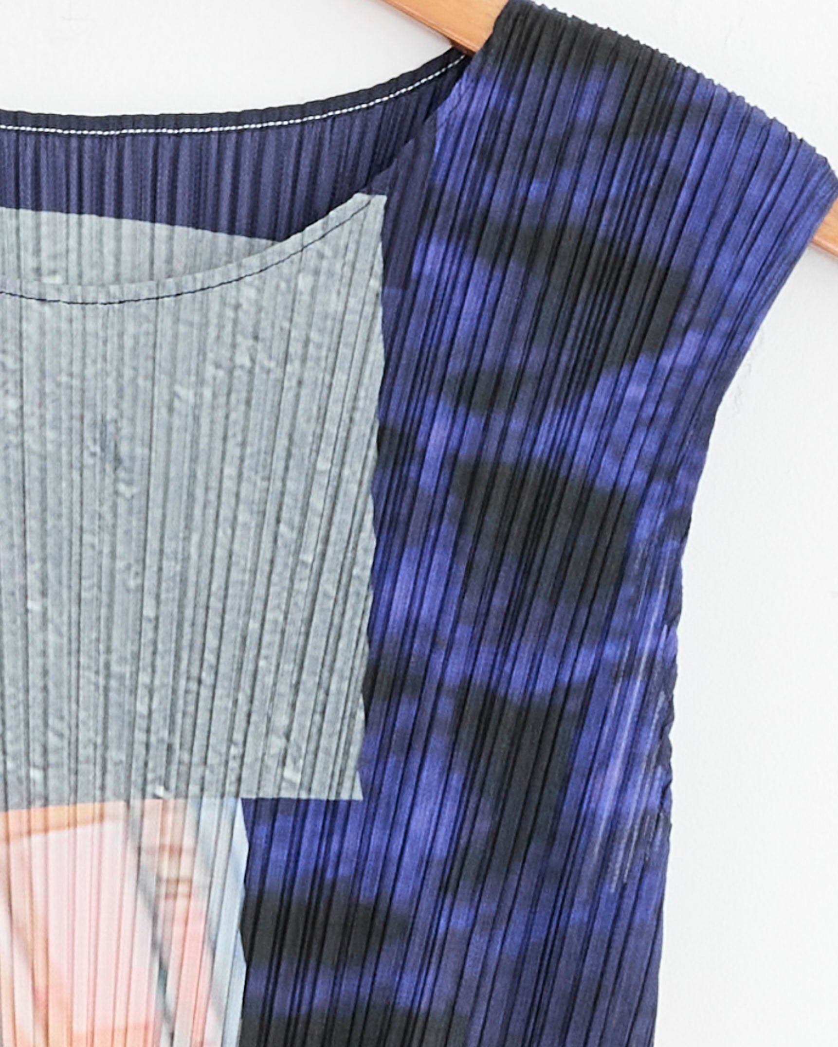 Close-up of the City Collage Dress in Grey/Navy by Pleats Please Issey Miyake—a pleated sleeveless dress with abstract blue, black, gray, and peach patterns—hanging on a wooden hanger against a white background.